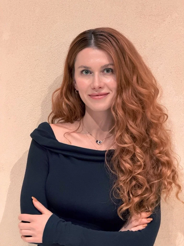 A woman with long, curly red hair, wearing a black off-shoulder top, standing against a beige wall.