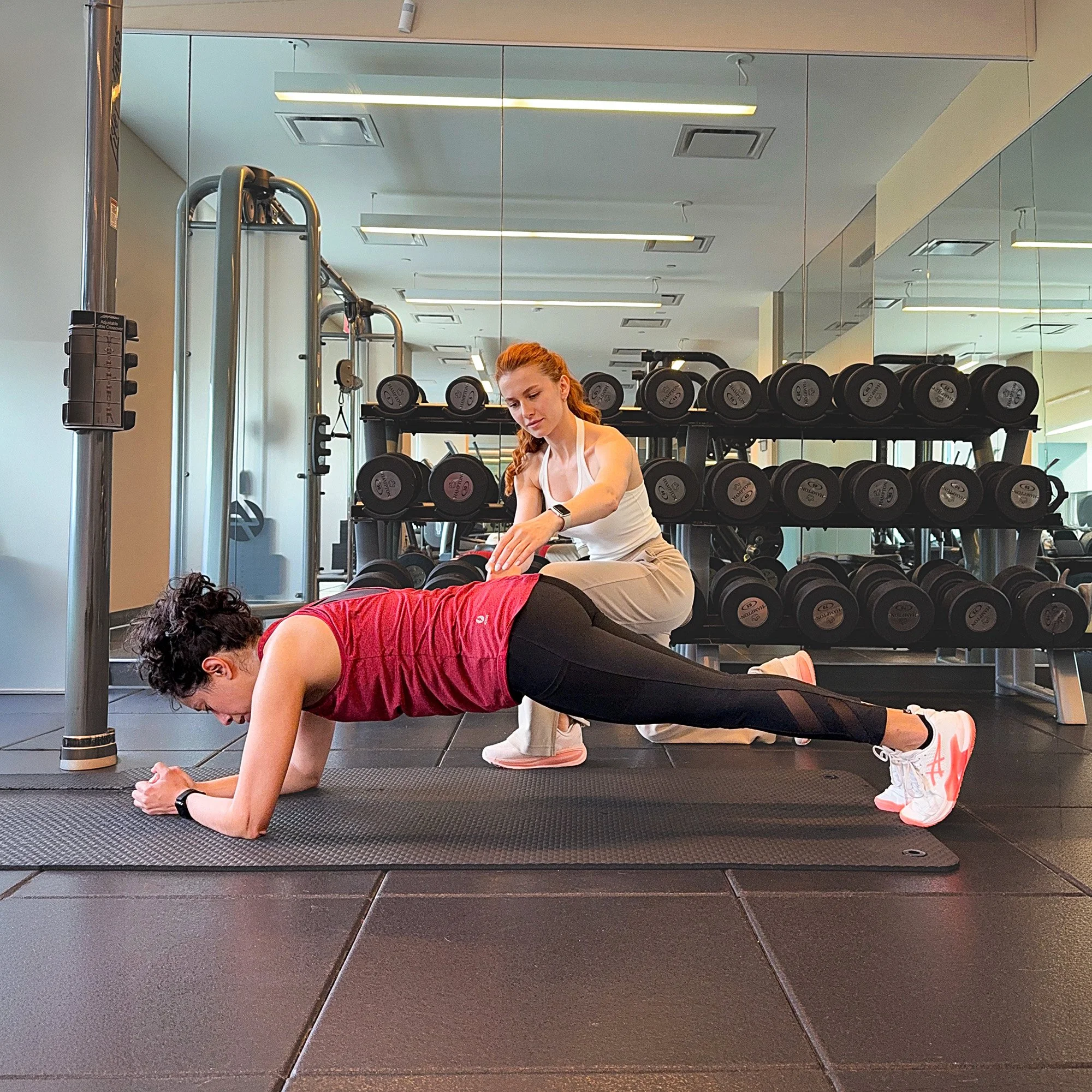 A woman in workout clothing performing a plank exercise on a yoga mat in a gym, with dumbbells and gym equipment visible in the background.