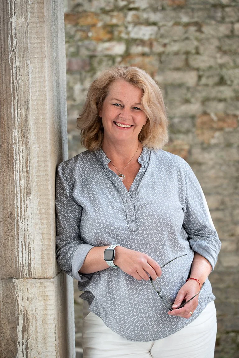 Rachel O'Connor, holding glasses and standing against a brick wall.