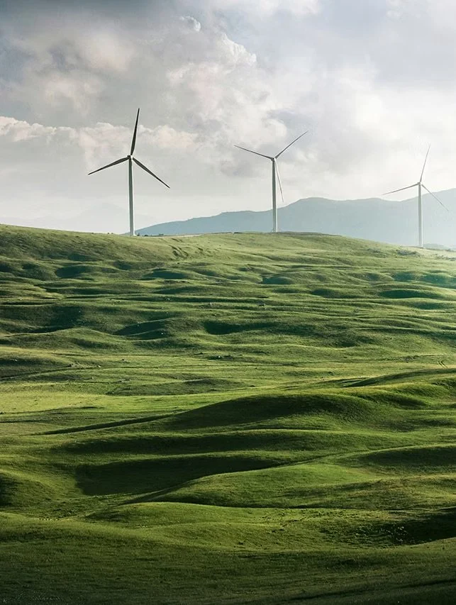 Green rolling hills with three wind turbines and a cloudy sky in the background.