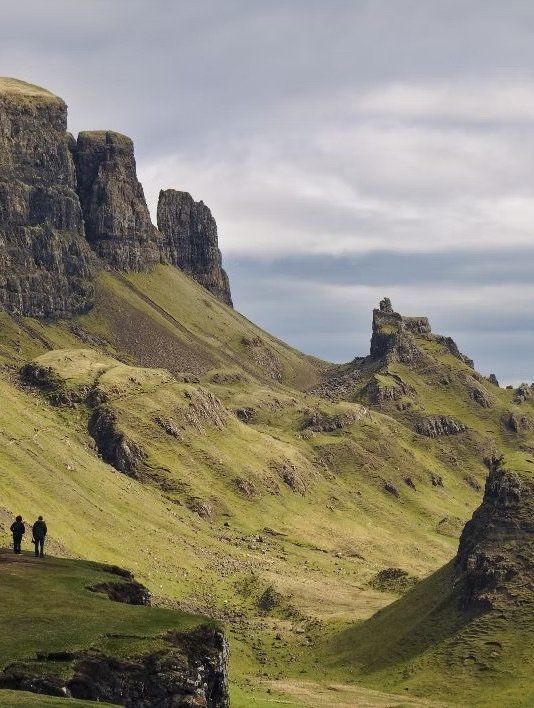 Two people walking on a grassy field in front of green cliffs and rocky formations under a cloudy sky.