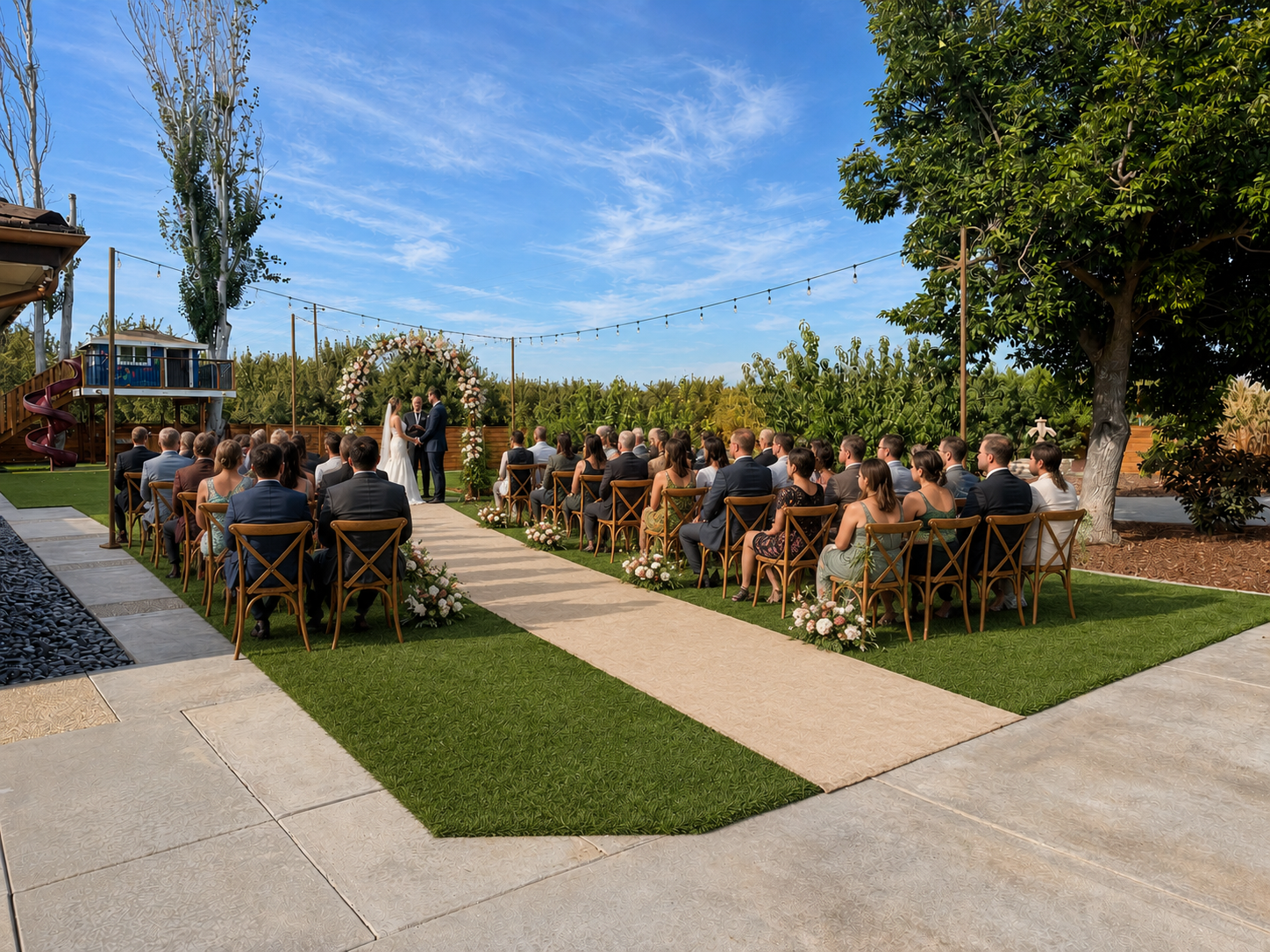 Outdoor wedding ceremony with guests seated on wooden chairs facing an officiant and bride and groom under a floral arch, on a sunny day with blue sky and lush greenery in the background.