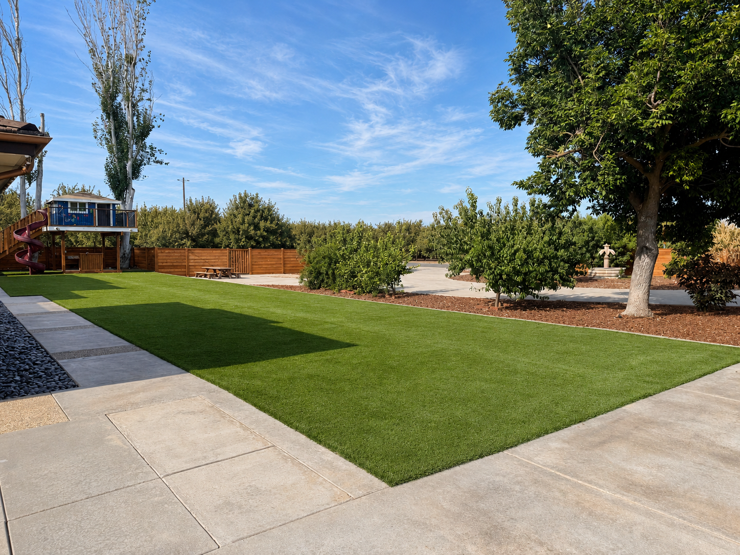 A backyard with a well-maintained lawn, concrete patio, trees, and a wooden fence under a blue sky with some clouds.