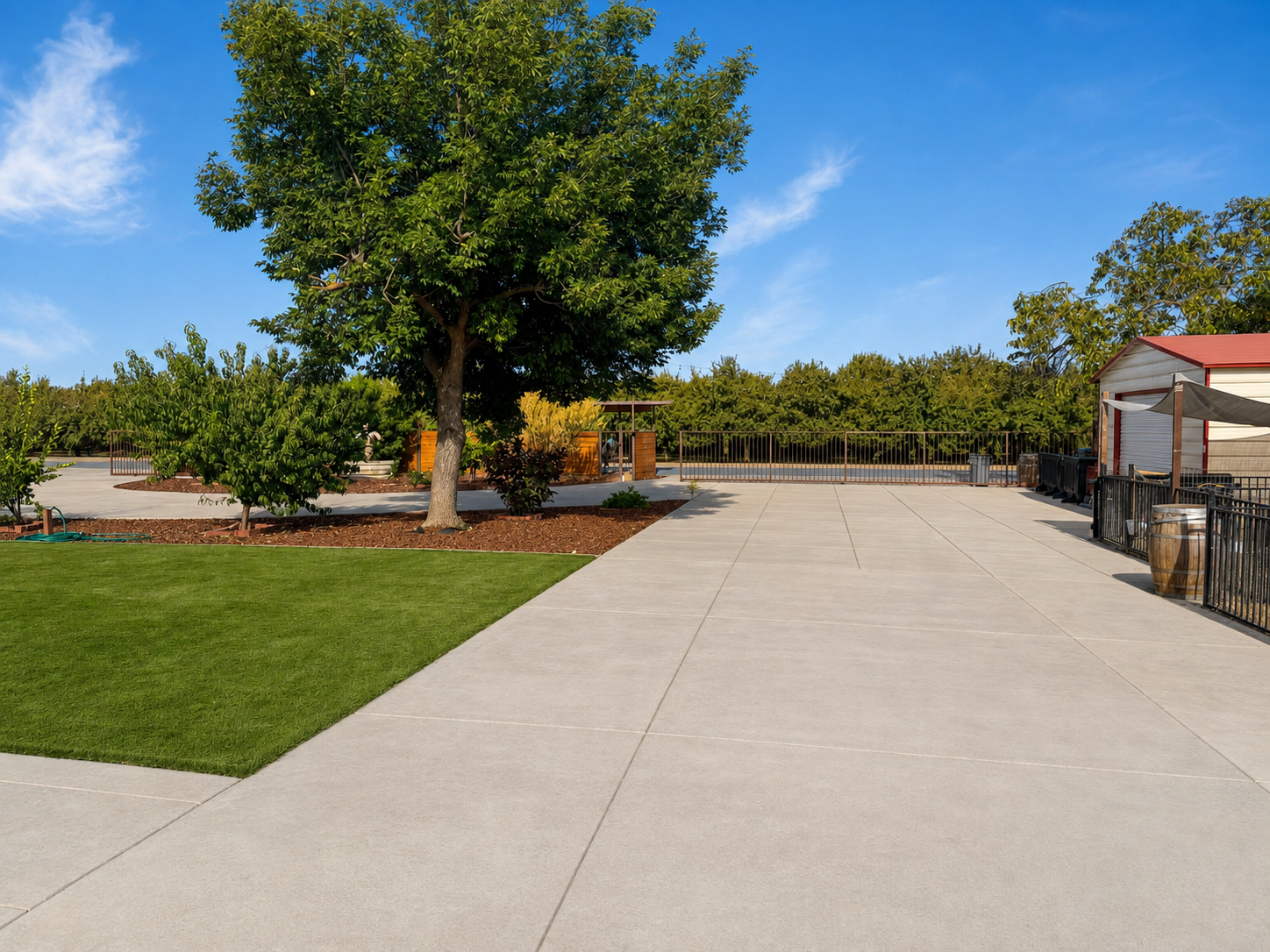A spacious outdoor patio area with concrete flooring, green grass, a large tree, and a clear blue sky. There are some smaller trees and plants, a red and white building with a shaded area, black metal fencing, and a wooden panel in the background.
