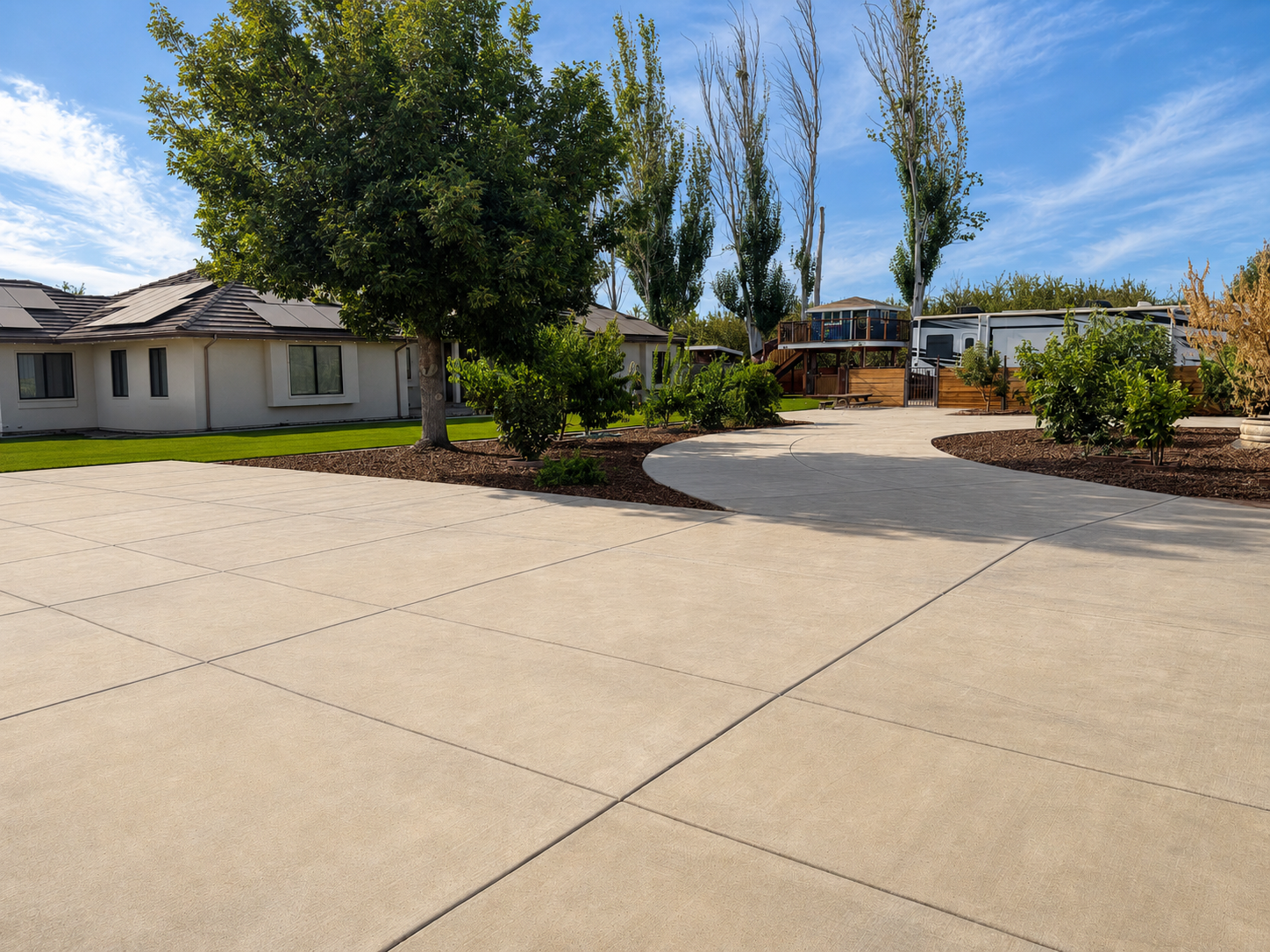 A spacious driveway curves around a landscaped yard with trees and bushes, leading to a house and a covered outdoor deck area in a suburban neighborhood under a partly cloudy sky.