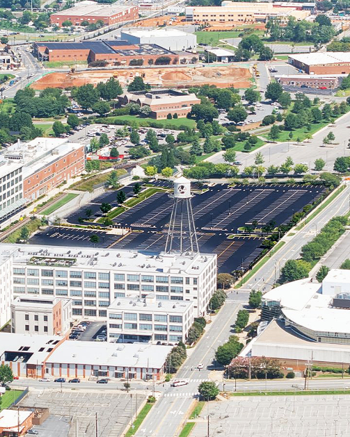 Aerial view of a cityscape featuring a white multi-story building with a water tower on top, black parking lot with many empty spaces, commercial and industrial buildings, green trees, and an under-construction area in the background.
