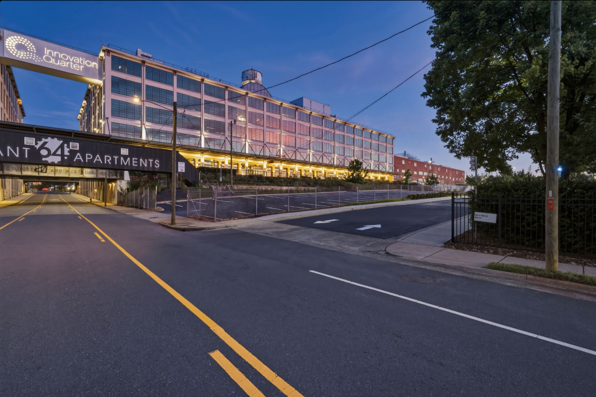 A city street scene at dusk showing a large industrial-style building with illuminated interior, a sign for 'Innovation Quarter,' a parking garage for apartments, and a street with yellow lines and a crosswalk, with trees and power lines in the background.