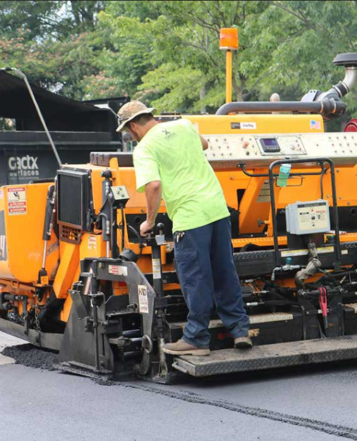 Worker operating a road paving machine on a freshly paved road, wearing a green shirt and a wide-brim hat.