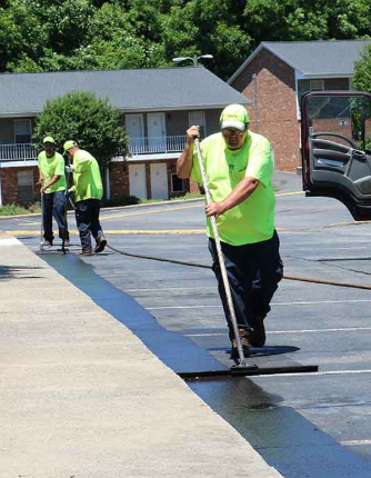 Workers in neon green shirts applying a black seal coating on a parking lot surface with brushes and hoses.