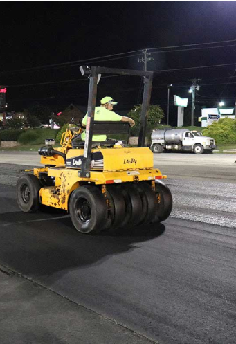 A construction worker operating a yellow steamroller at night on a street.