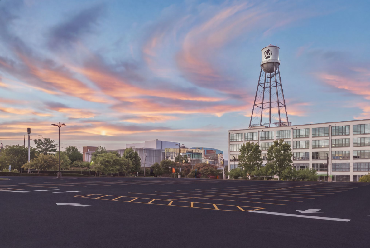 Empty parking lot with a large water tower marked with the number 64, modern buildings, trees, and a colorful sunset sky with pink and orange clouds.