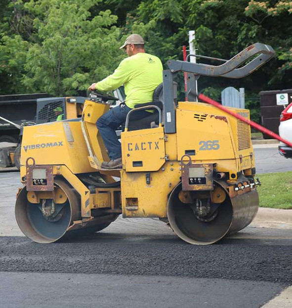 Worker operating a yellow Vibromax road roller to compact asphalt pavement on a street.