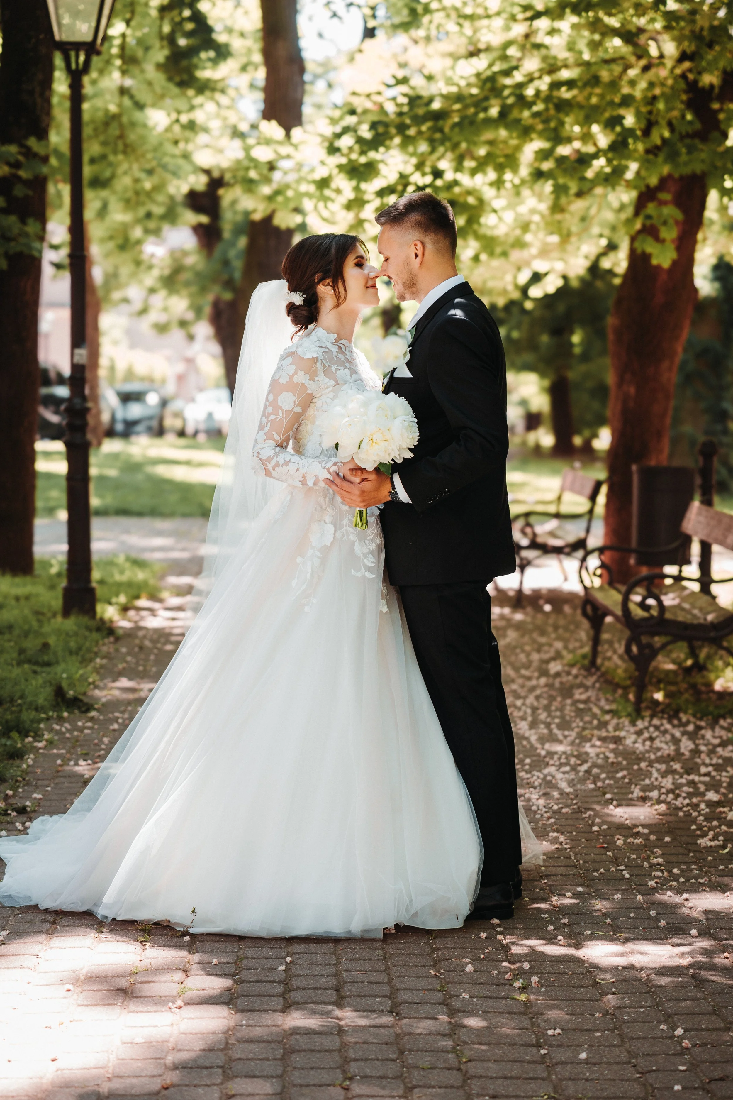 A bride and groom stand close together outdoors, touching noses, under sunlight filtering through trees, with benches and parked cars in the background, during their wedding.