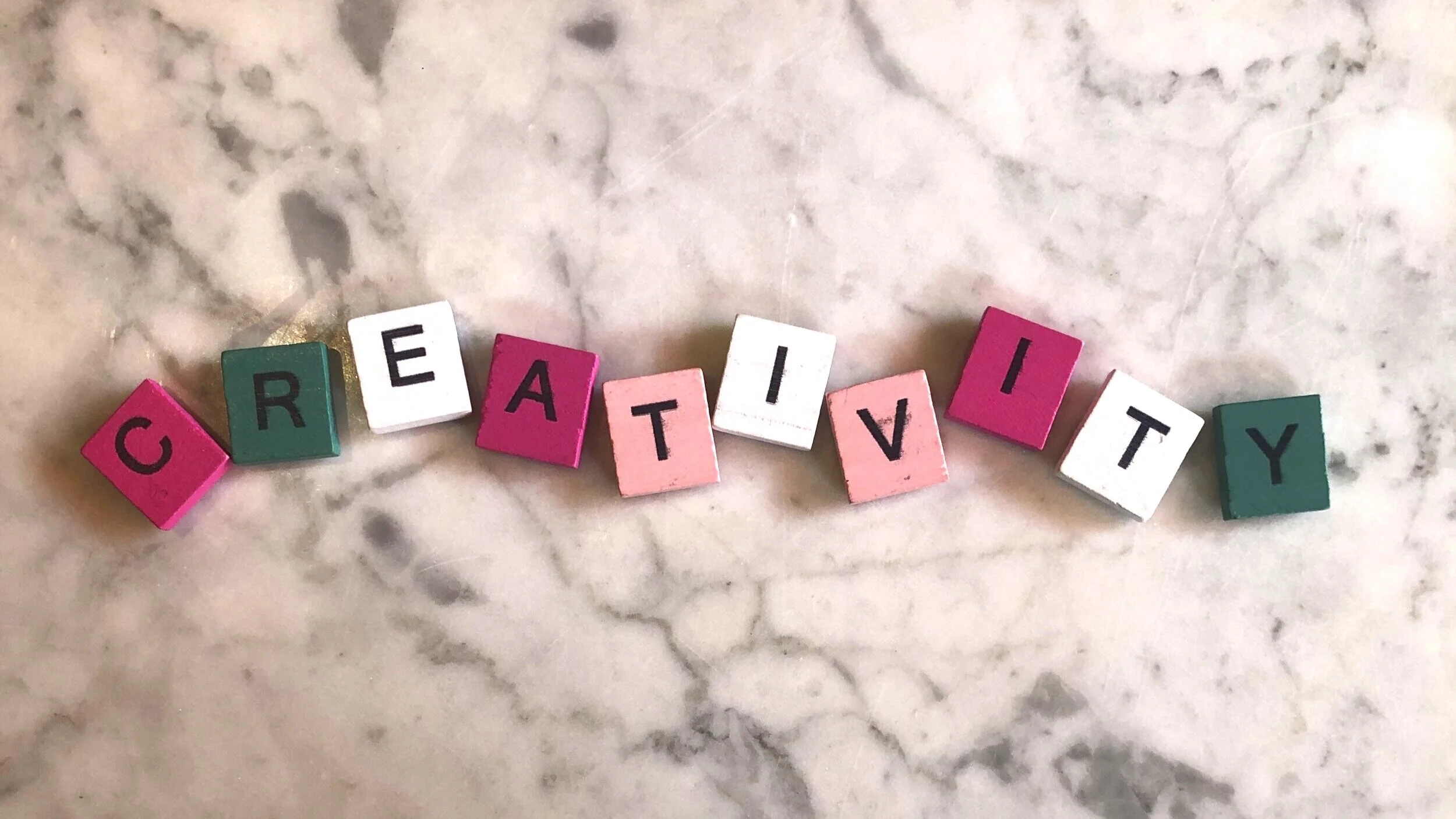 Scrabble letters in pink, green and white spelling our CREATIVITY on a marble surface