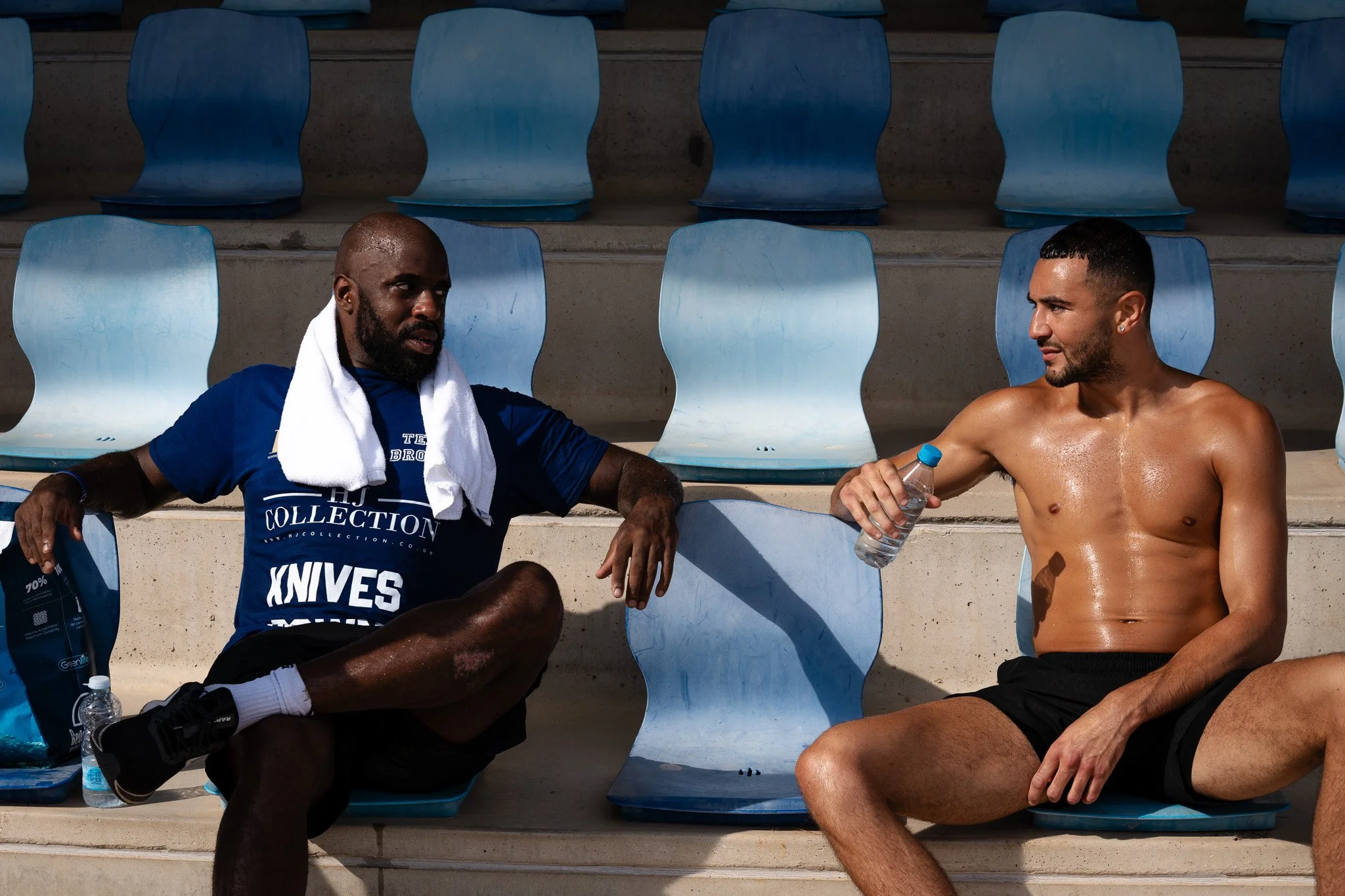 Two shirtless men sitting on blue stadium seats, one with a white towel around his neck, having a conversation after exercise. One man is holding a bottle of water, and there are water bottles and a bag nearby.