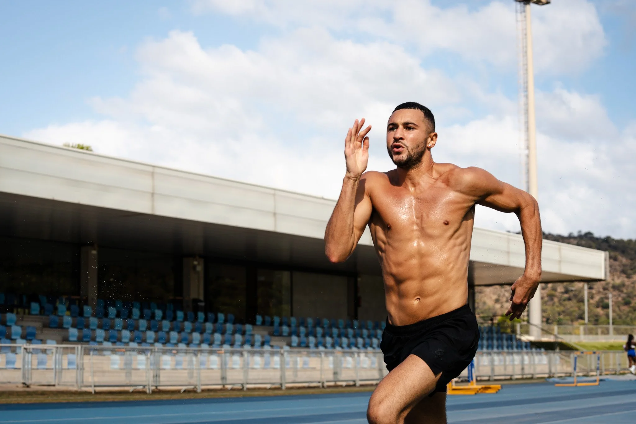 A shirtless man running on an outdoor track, sweating, with an empty stadium and blue sky in the background.