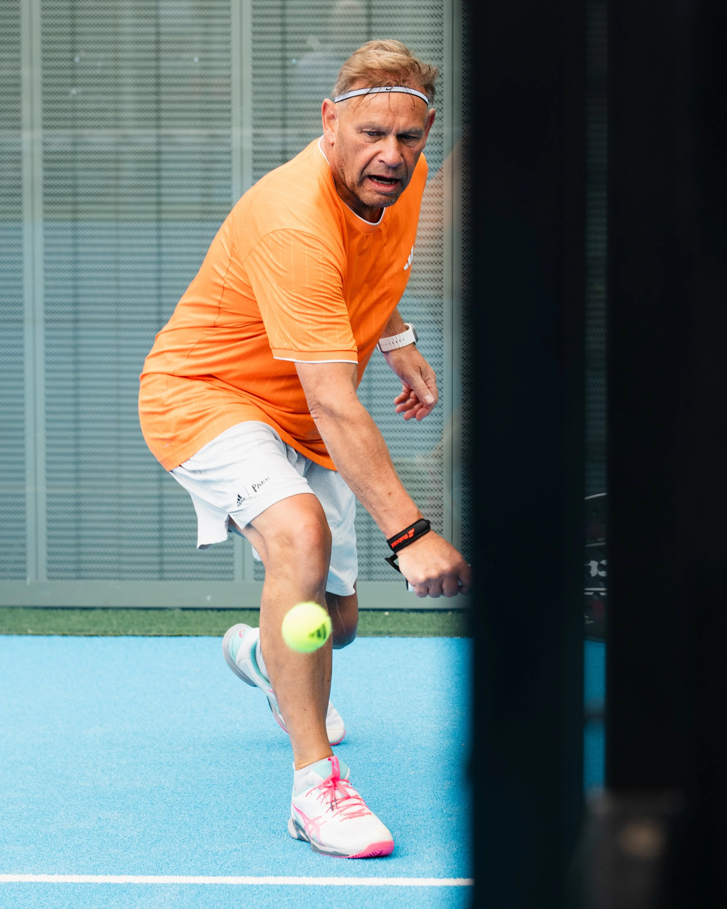A man in an orange shirt, white shorts, and white and pink sneakers is playing tennis on a court. He is bending down to hit a tennis ball with his right hand, wearing a white wristwatch and a sweatband.