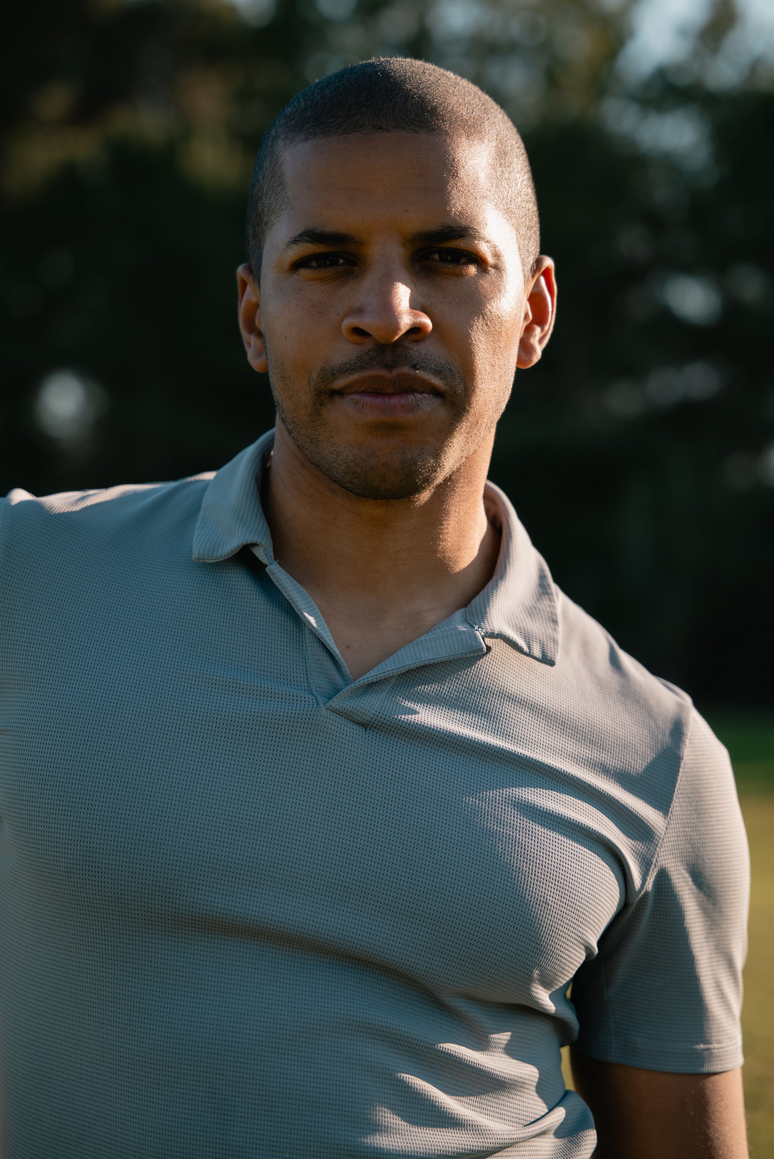 Close-up of a man with short black hair and a beard, wearing a light gray collared shirt, standing outdoors with blurred trees in the background, illuminated by sunlight.