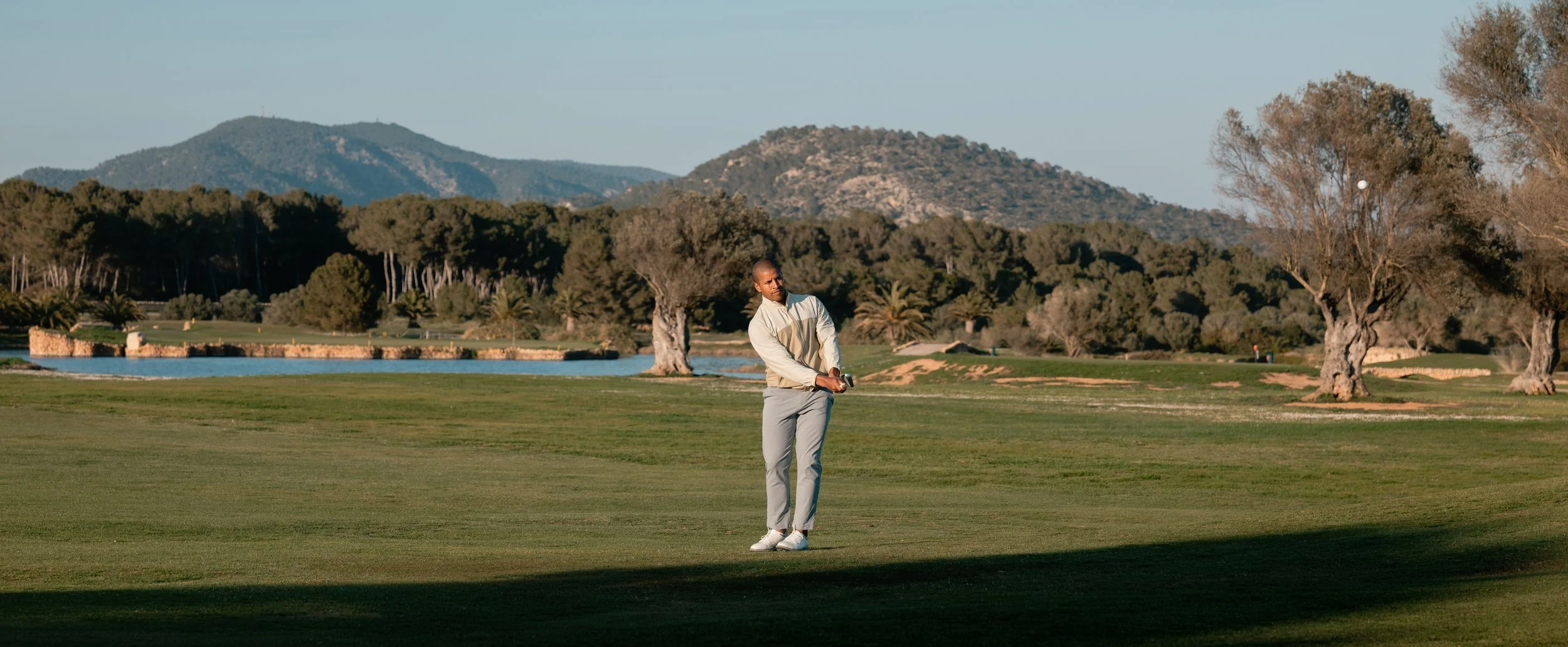 A man on a golf course preparing to hit a golf ball, surrounded by trees and mountains in the background during daytime.