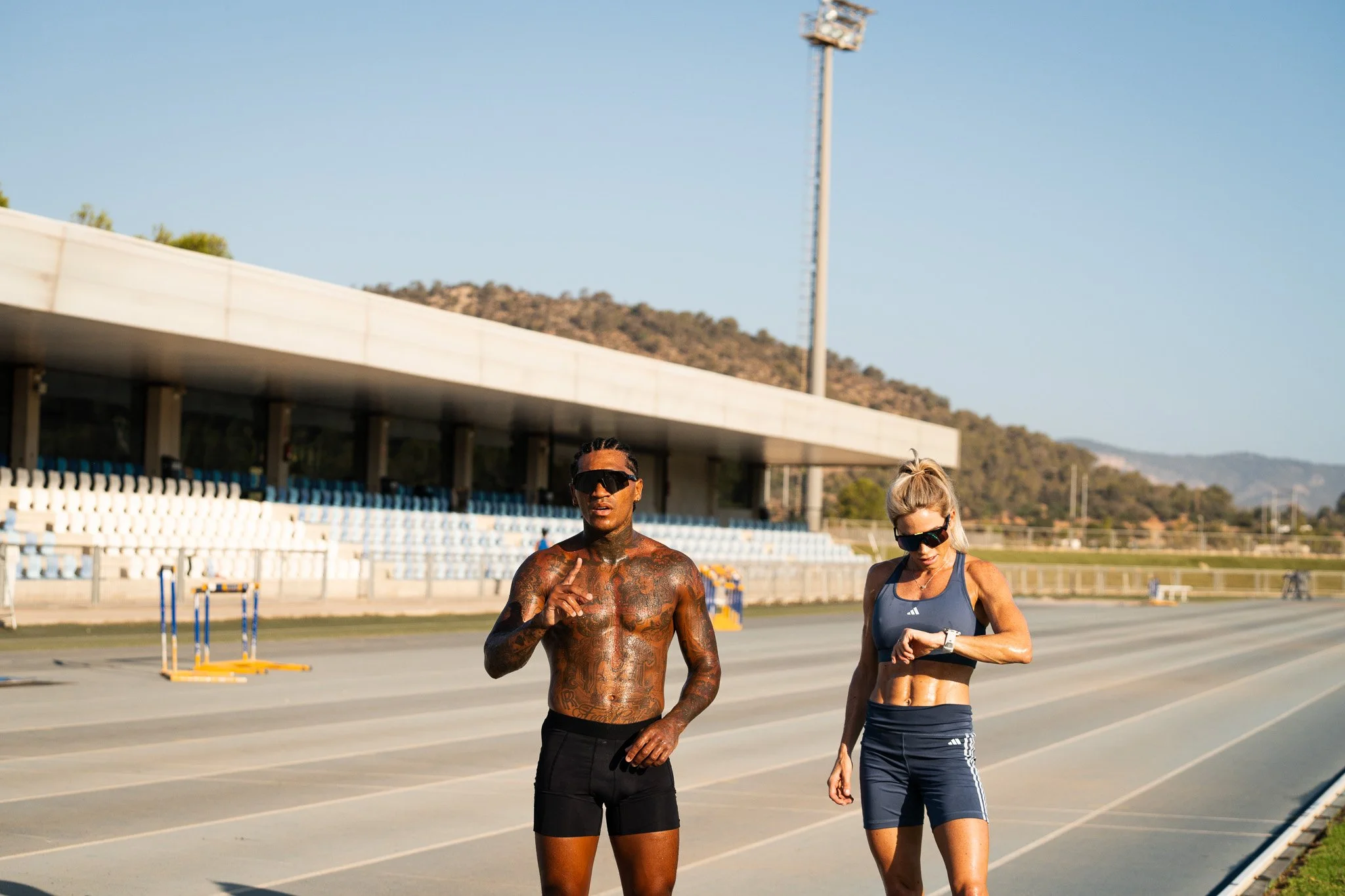 Two athletes, a shirtless man with tattoos and a woman in a sports bra, run on an outdoor track.