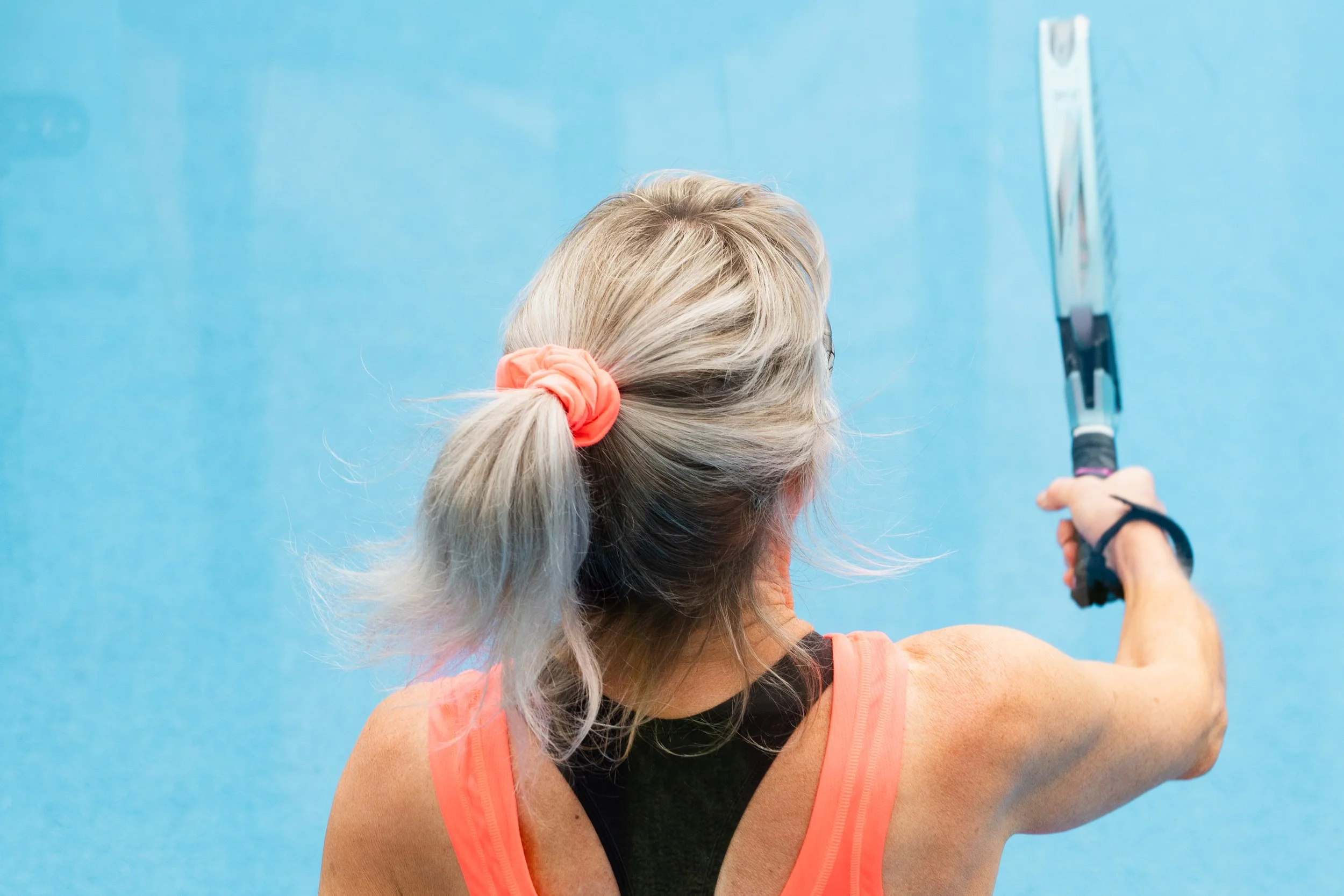 A woman with blonde hair tied back with a coral hair scrunchie is reaching up on a tennis court with a racket in her hand.