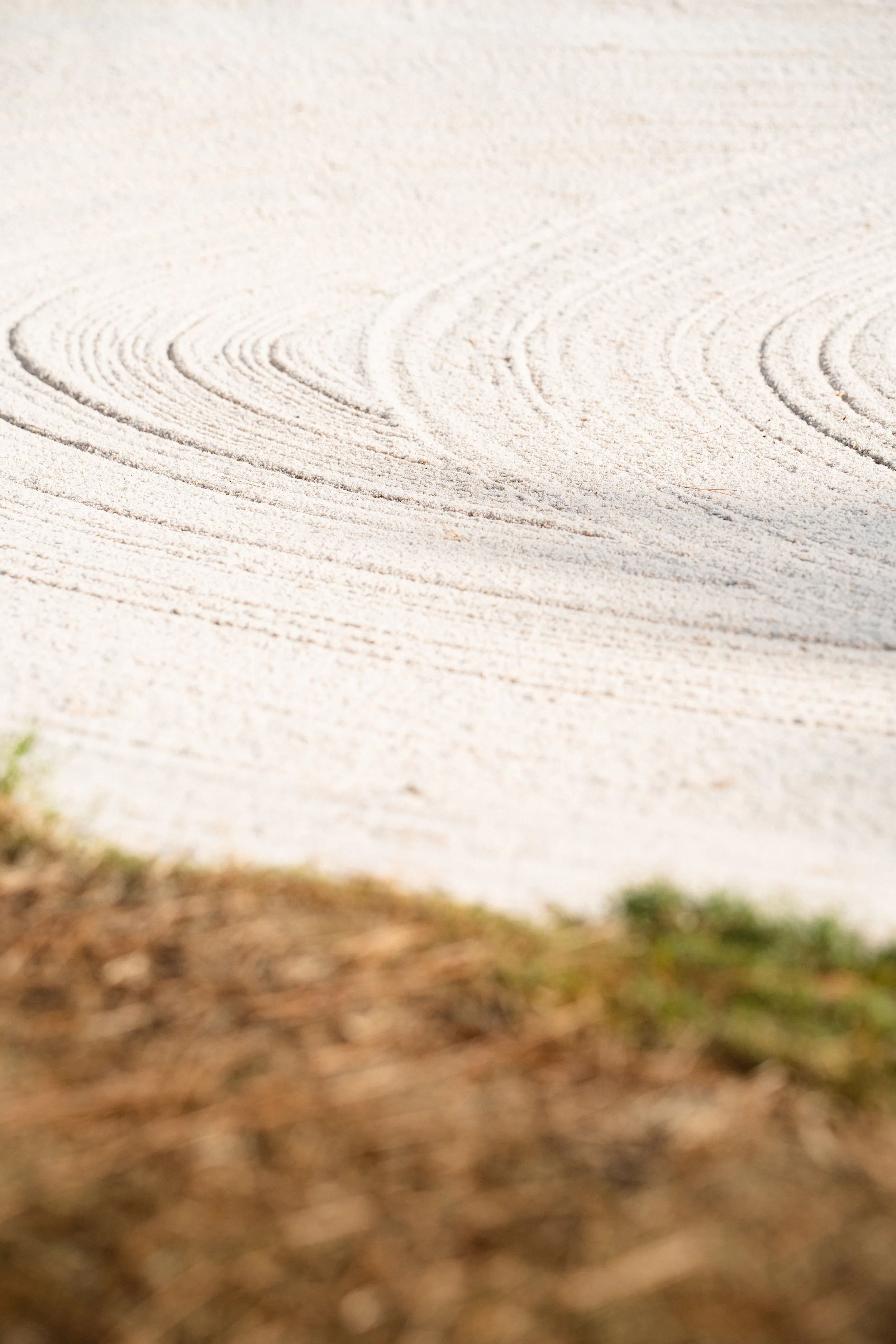 Close-up of raked white sand with circular patterns at a Japanese Zen garden, foreground showing blurred brown and green grass.