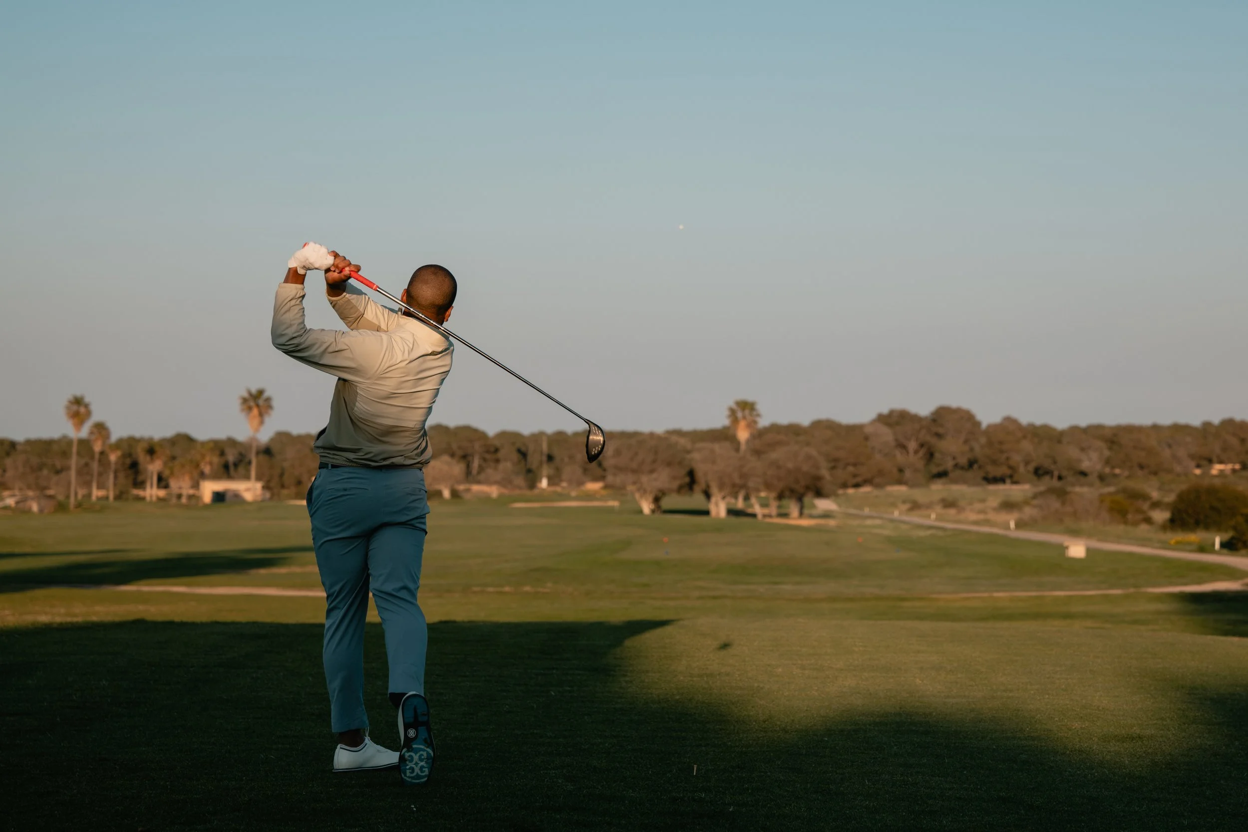 A golfer swinging a club on a golf course at sunset, with trees and a clear sky in the background.
