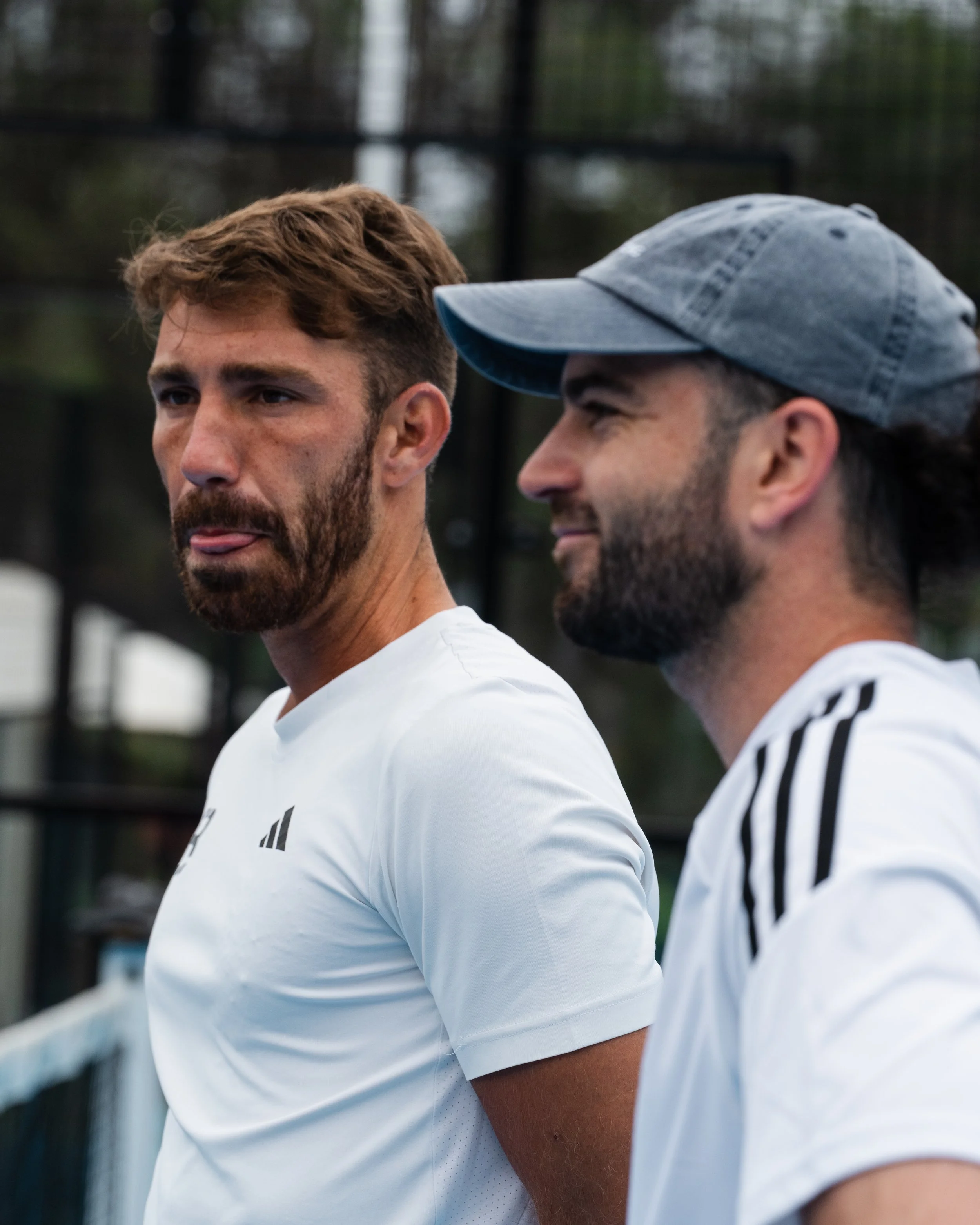 Two young men with beards and athletic attire standing outdoors, one with a cap, against a blurred background of trees and a black fence, during daytime.