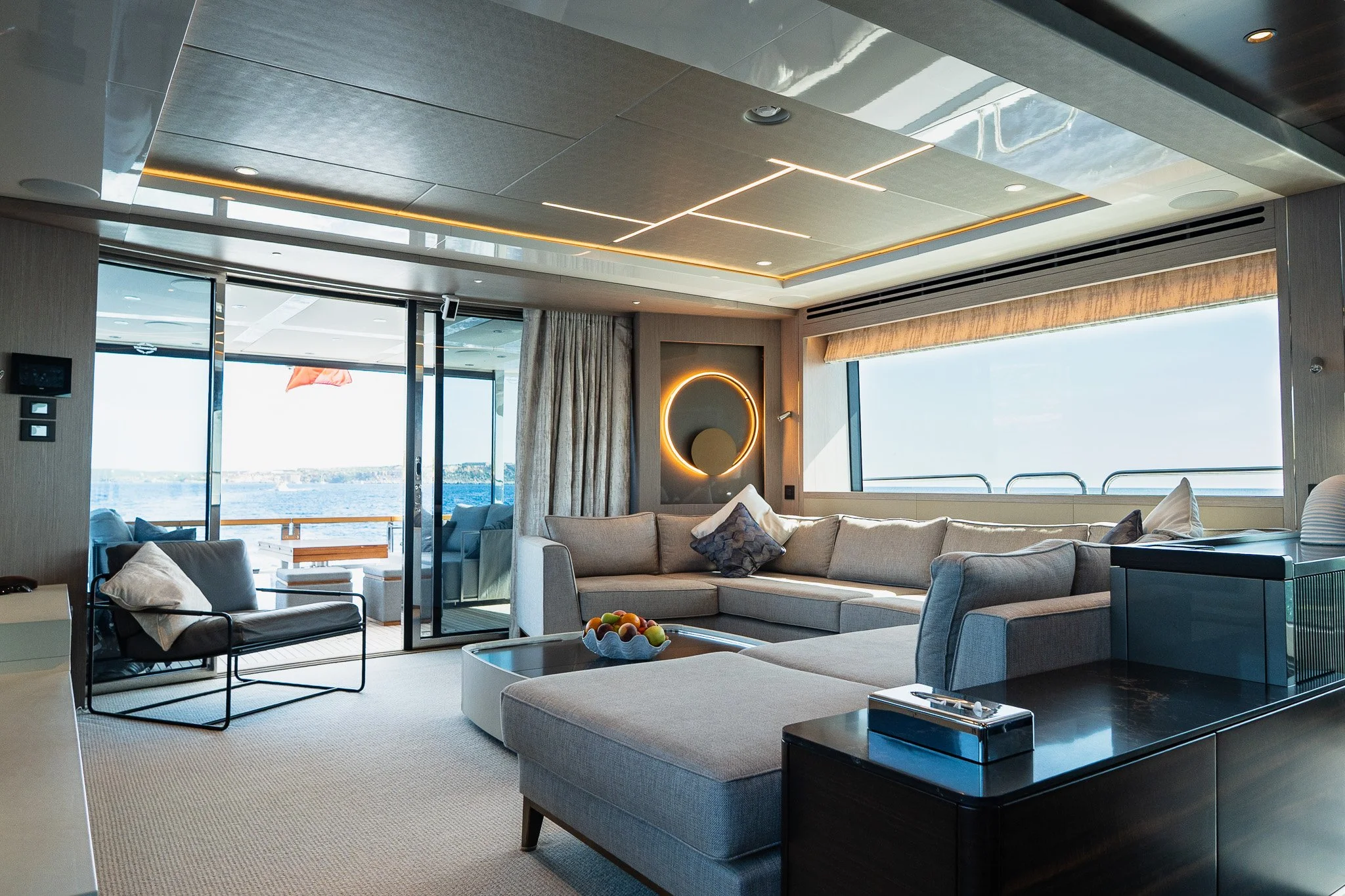 Modern living room with large sofa, armchair, coffee table with fruit, and floor-to-ceiling windows showing a view of water and sky outside.