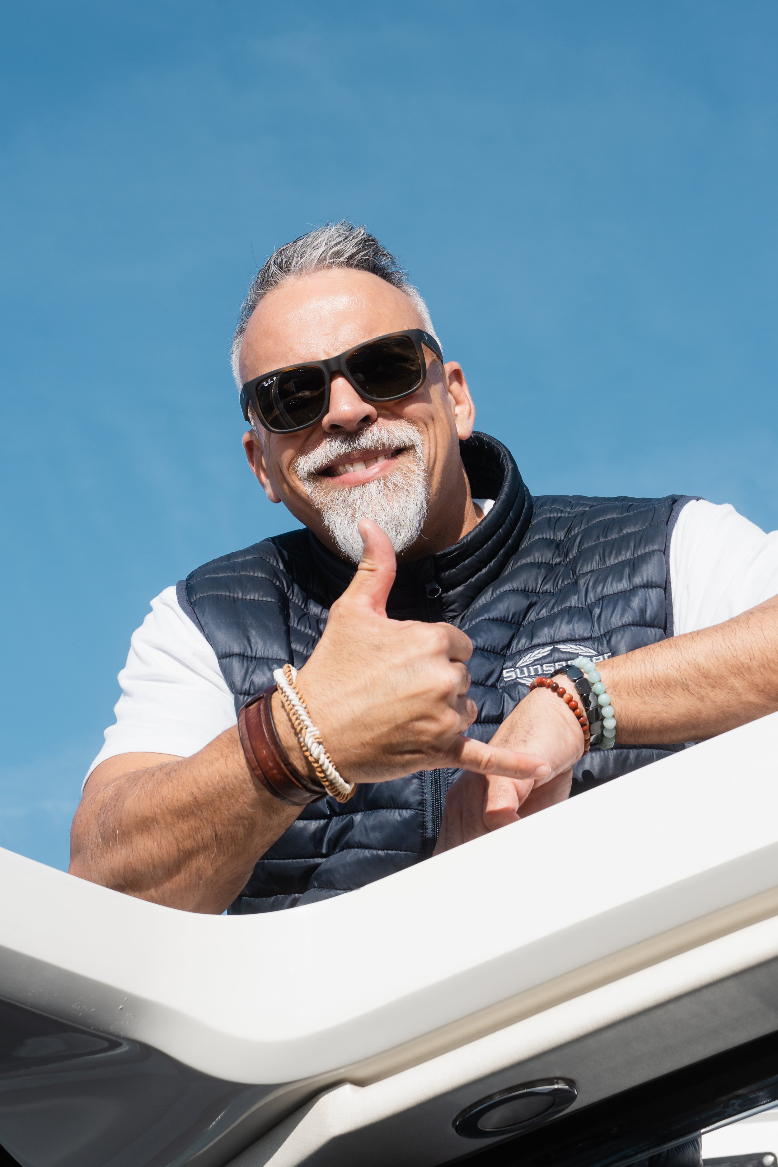 A smiling man with gray hair and a beard wearing black sunglasses, a black quilted vest over a white shirt, and multiple bracelets on his wrist giving a thumbs-up gesture against a clear blue sky.