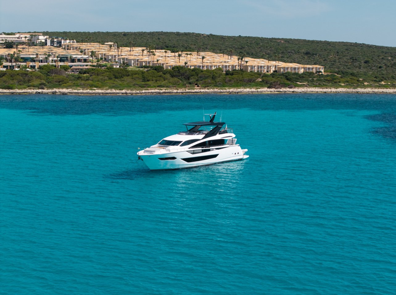 A white yacht on clear turquoise water near a rocky shoreline with green vegetation and a hillside residential area with beige buildings in the background.