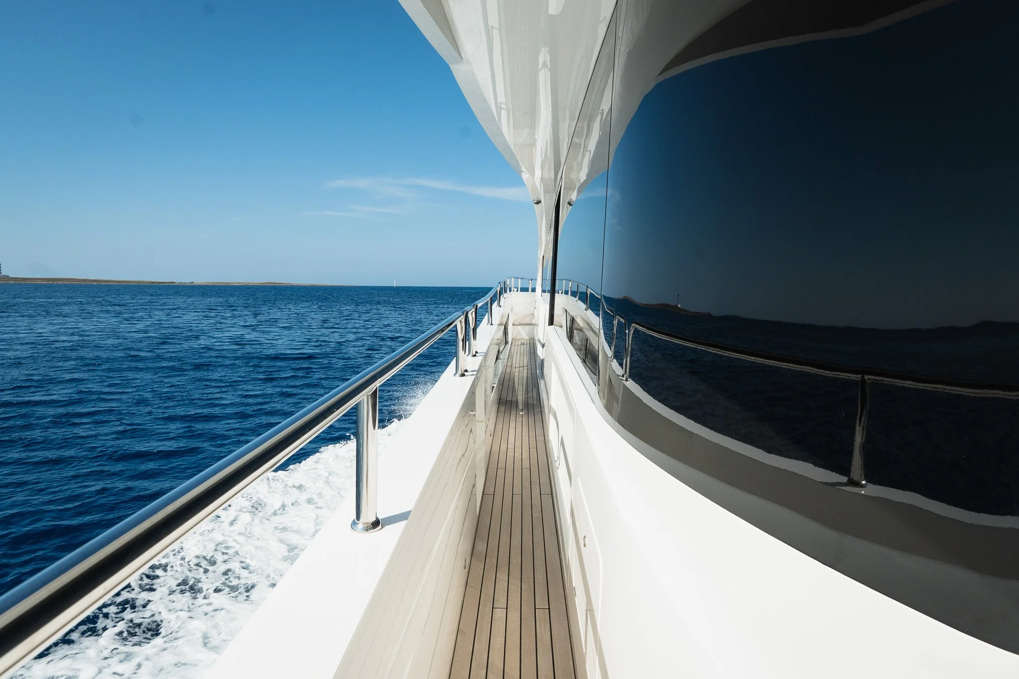 Side view of a white yacht sailing on the ocean with a clear blue sky, a distant shoreline, and a small sailboat on the horizon.