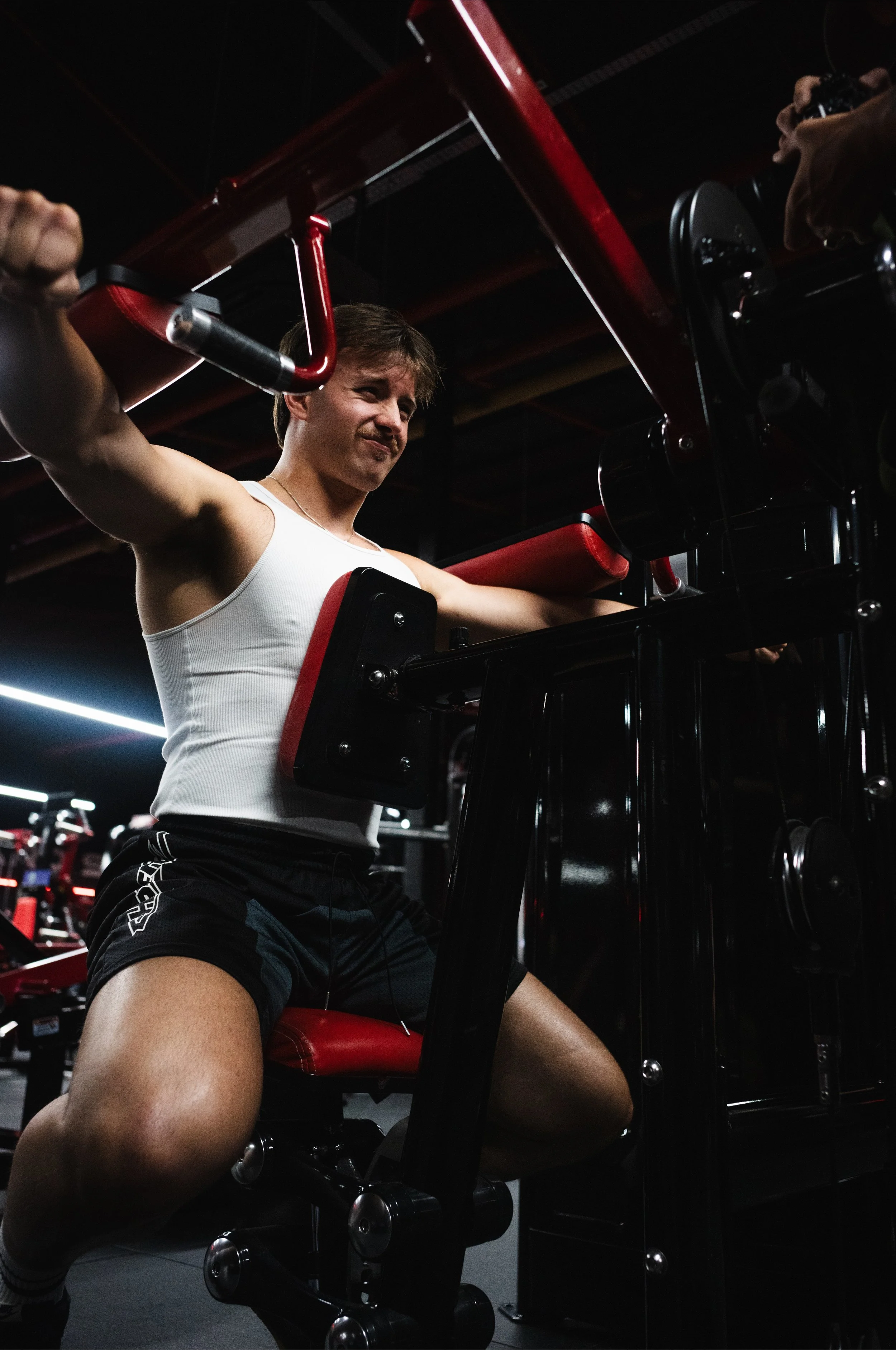 A man working out on a chest press machine in a dark gym, wearing a white tank top and black shorts.