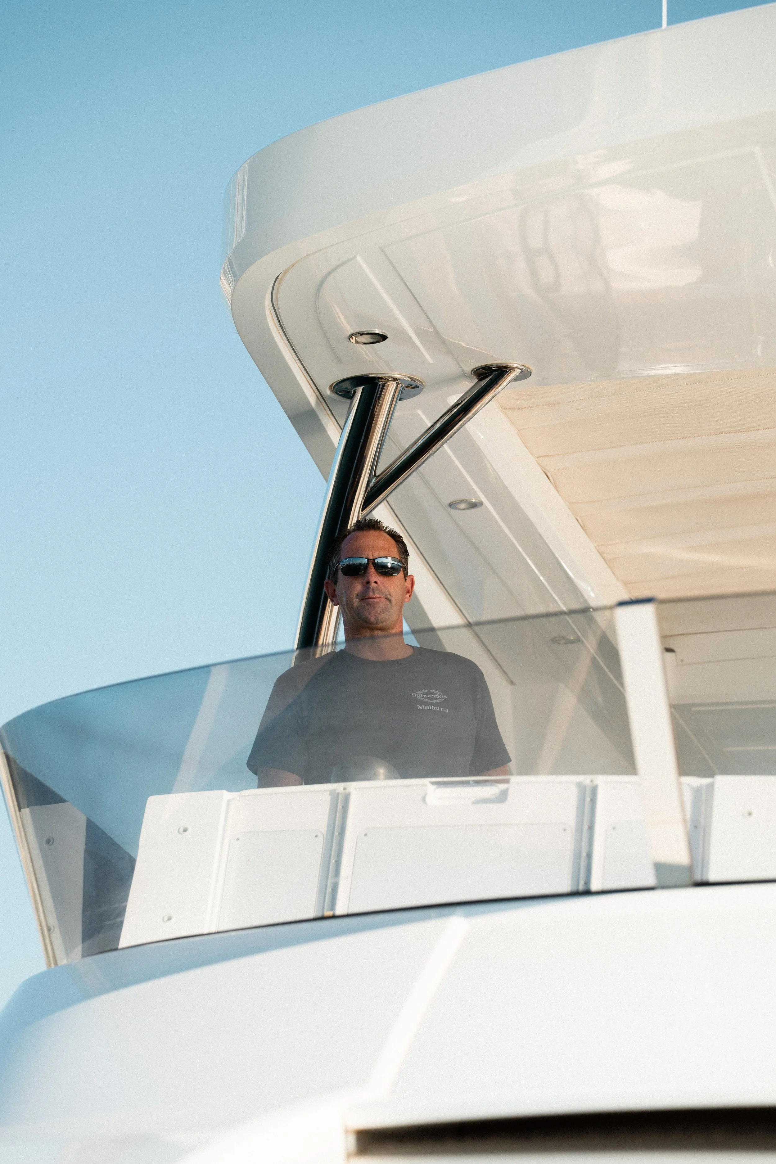 A man wearing sunglasses and a black t-shirt standing on a boat's balcony, with the clear blue sky in the background.