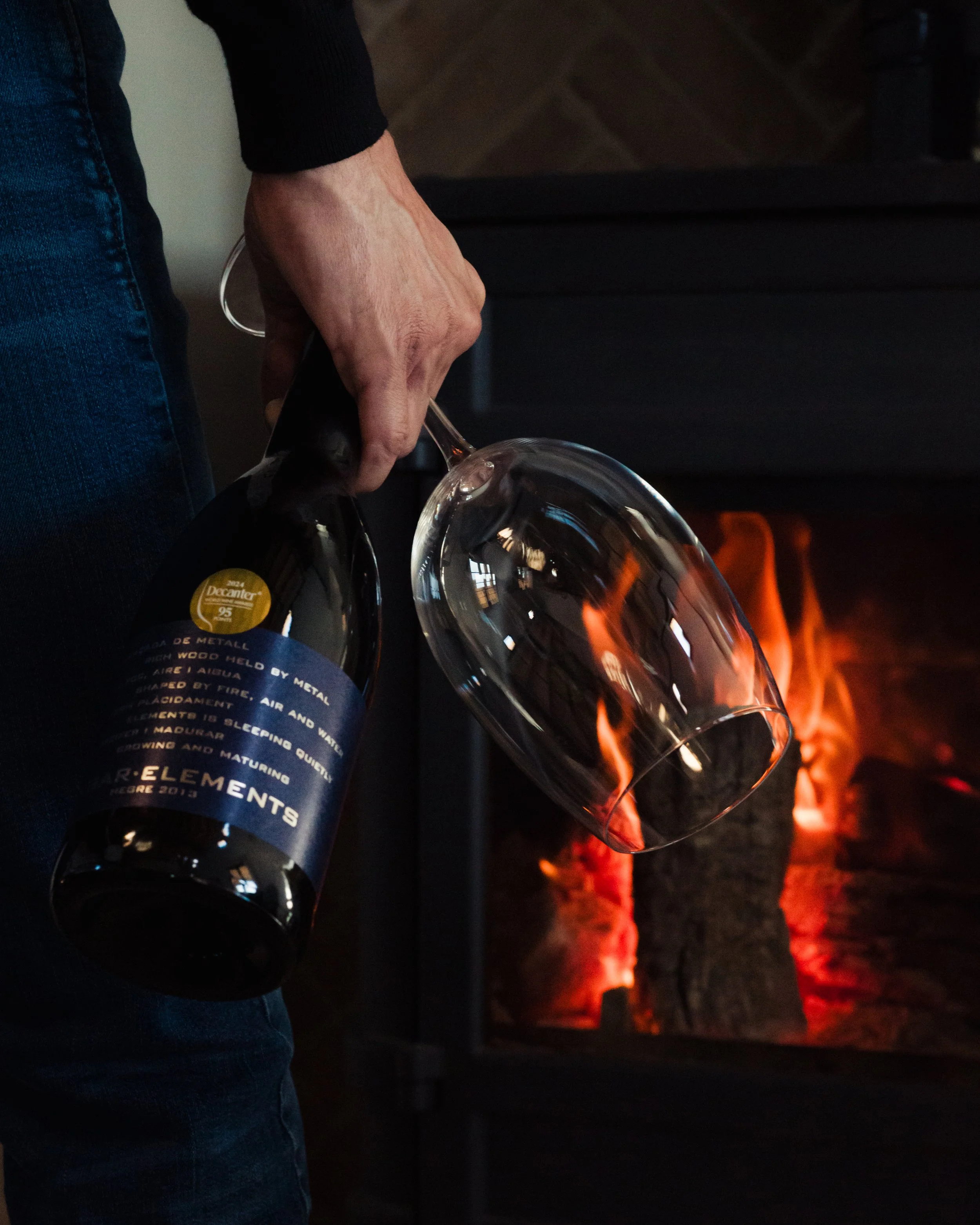 Person holding a glass of red wine near a fireplace with burning logs.