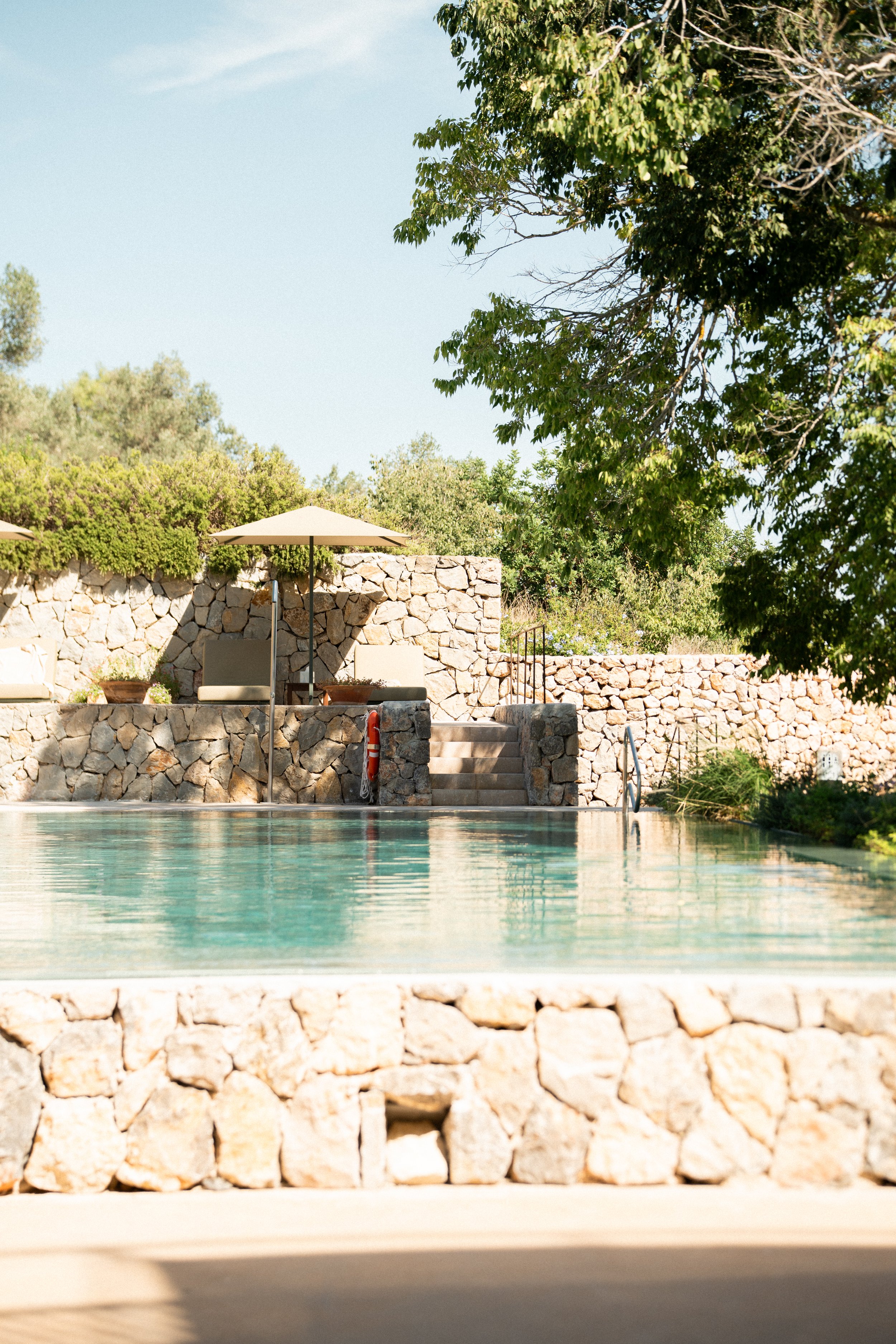A serene outdoor swimming pool surrounded by a stone wall and lush green trees, with poolside lounge chairs and umbrellas.