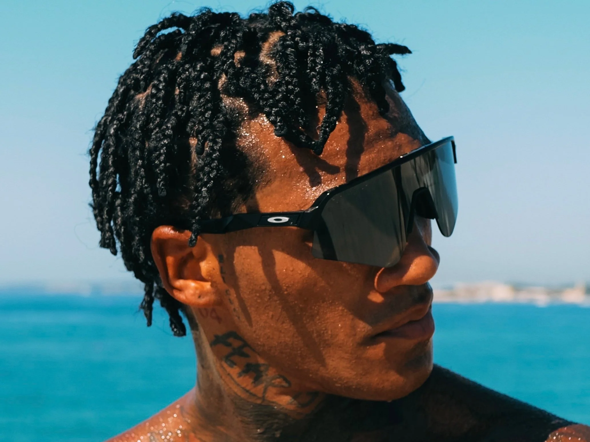 Person wearing black sunglasses with short twists hairstyle, standing by the ocean.