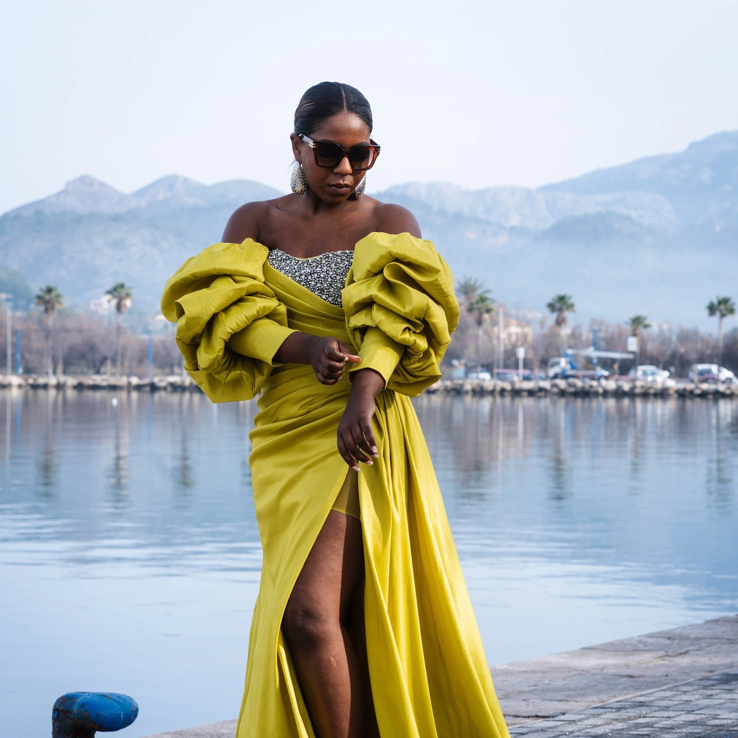 Person wearing a yellow dress with dramatic sleeves and sunglasses standing near a waterfront, with mountains and palm trees in the background.