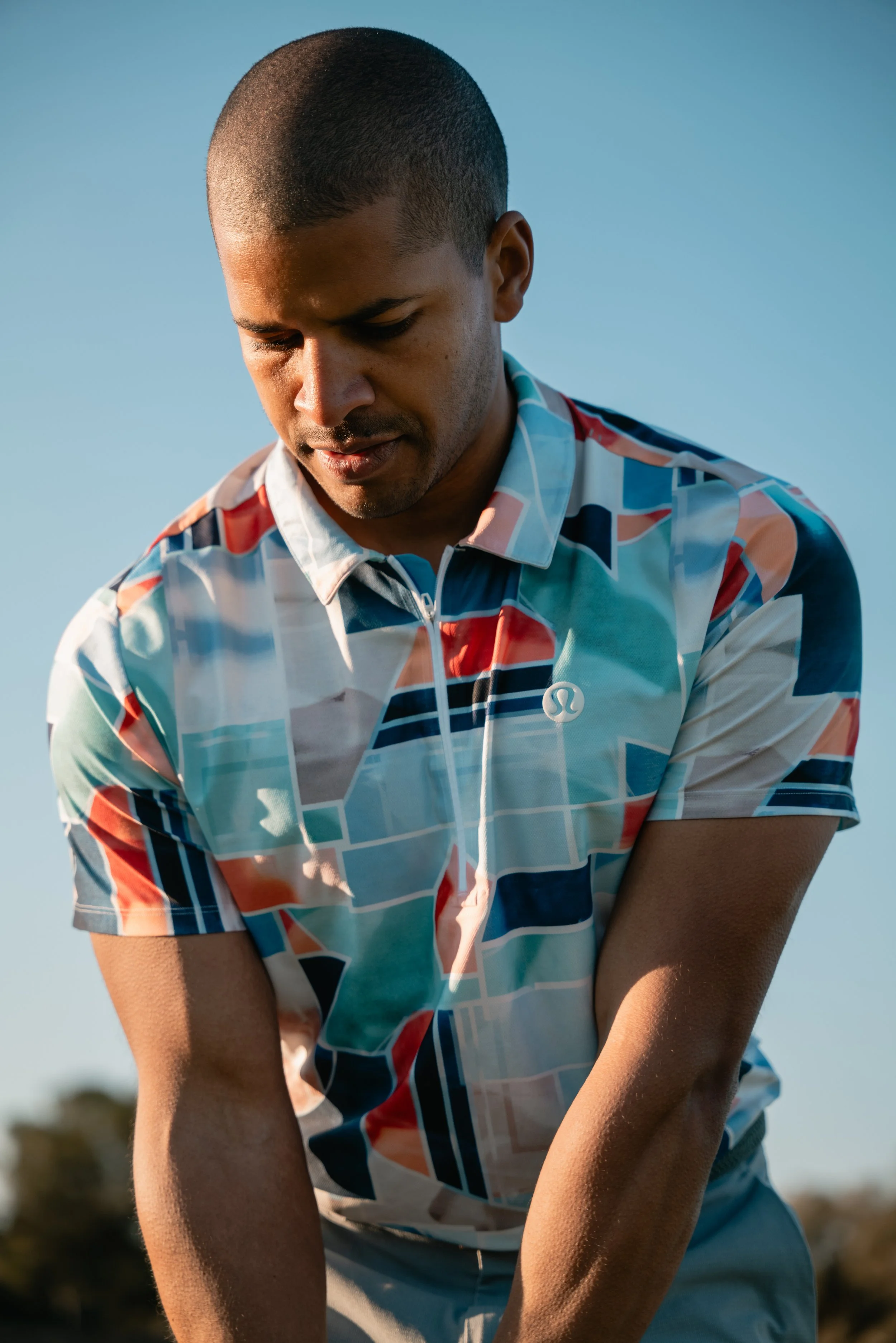 A man with short hair wearing a colorful, geometric patterned shirt outdoors with a blue sky in the background.