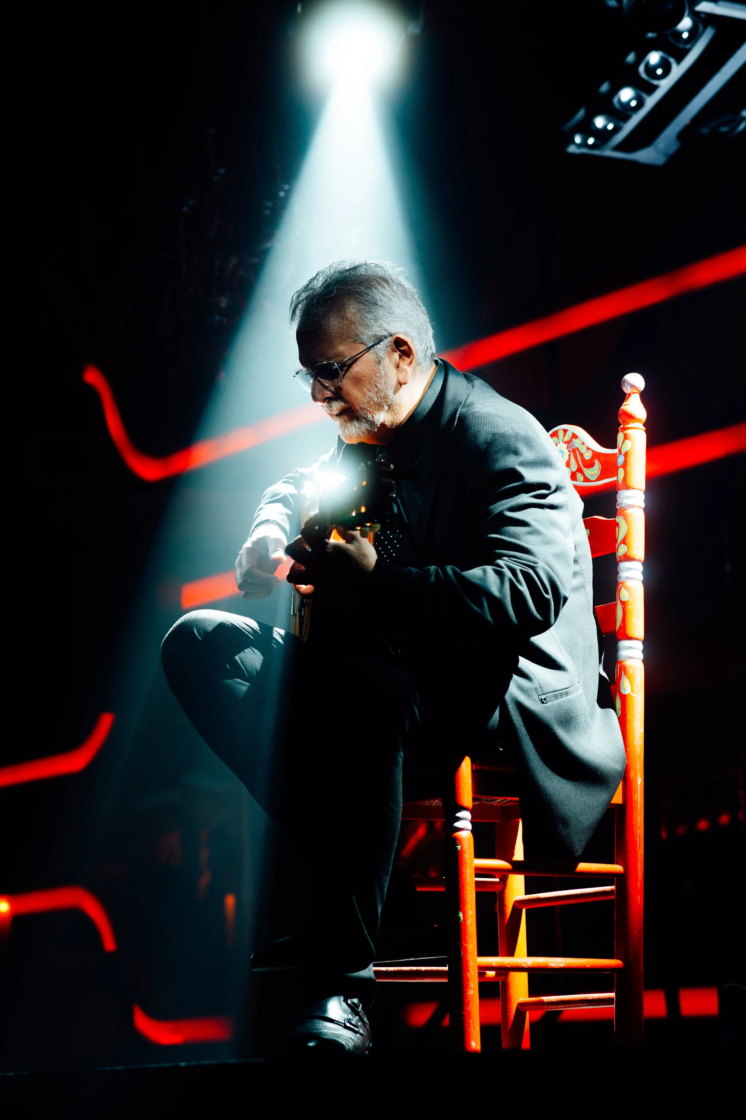 Man playing guitar on stage under spotlight with red chair and dramatic lighting.