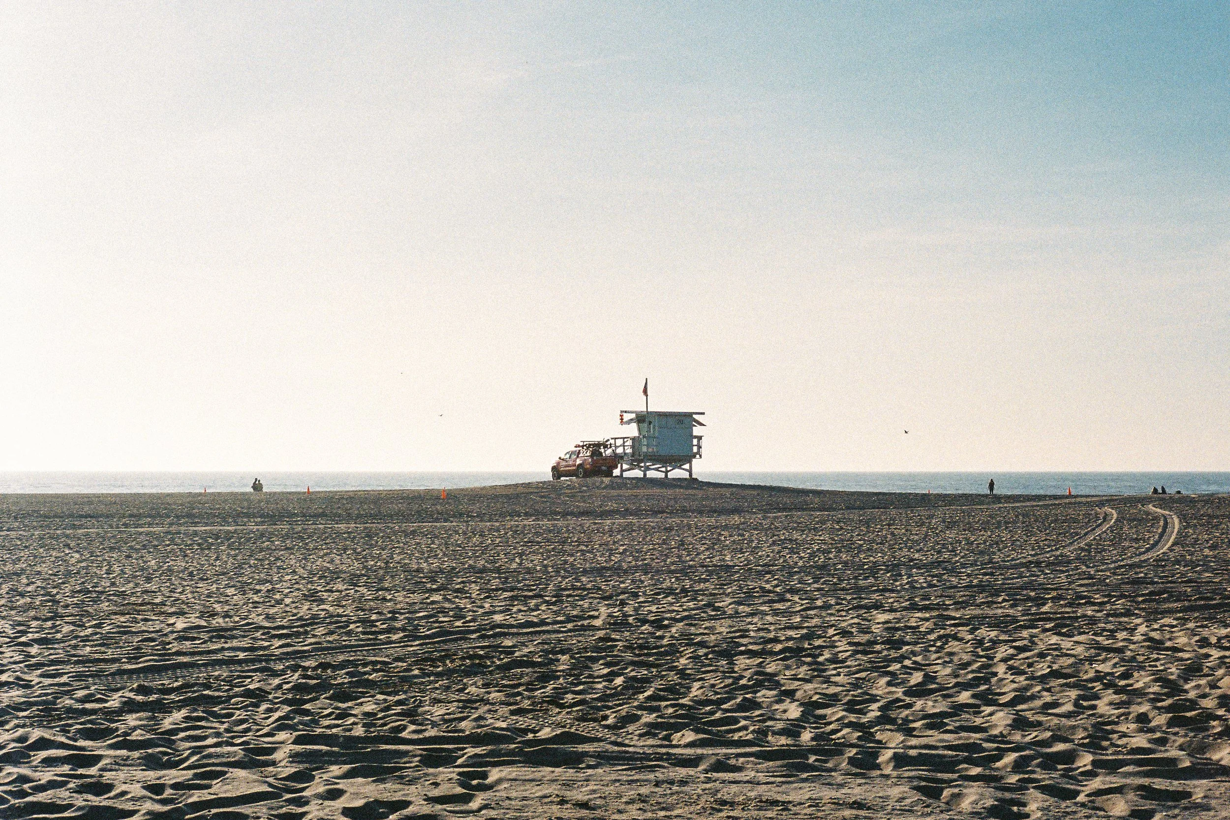A lifeguard tower on a sandy beach with a pickup truck parked beside it, and a few people in the distance near the shoreline, under a clear sky.