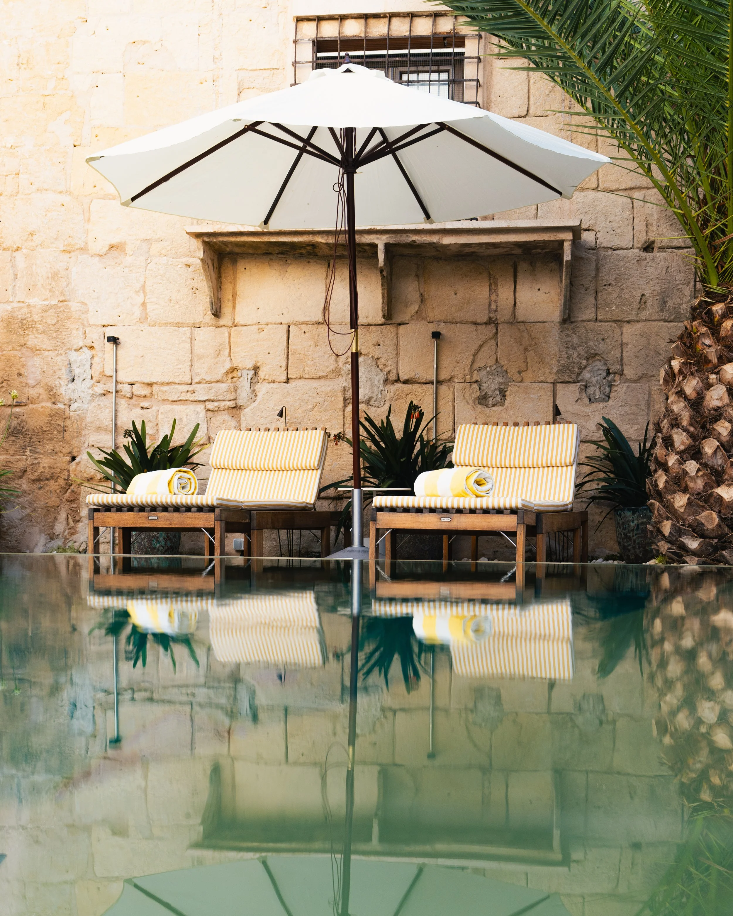 Poolside scene with two yellow and white striped lounge chairs under a large white umbrella, with rolled towels on each chair. There are two potted plants behind the chairs, a stone wall with a window with bars, and part of a palm tree on the right s