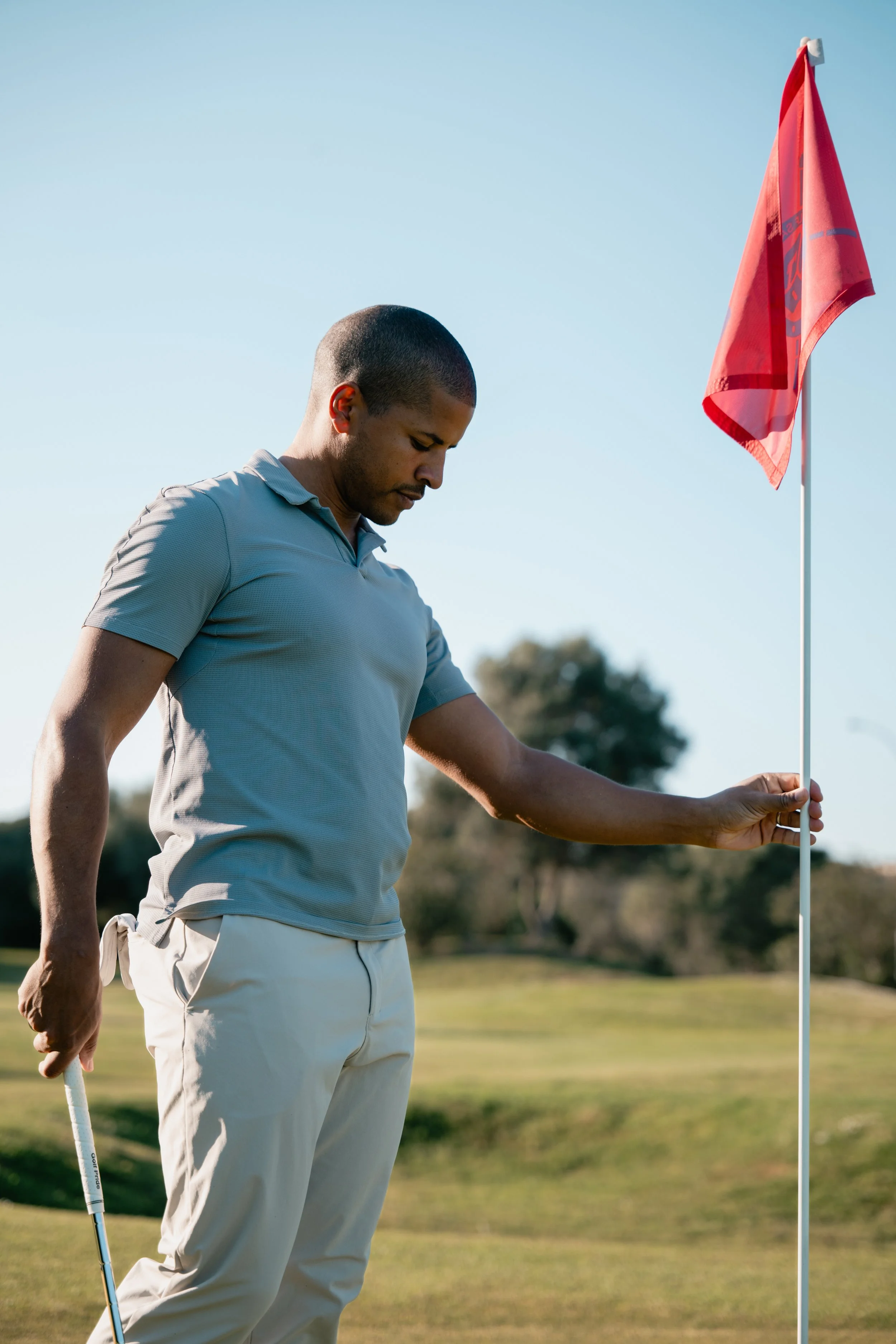 A man with short hair wearing a gray polo shirt and white pants standing on a golf course holding a golf flag.