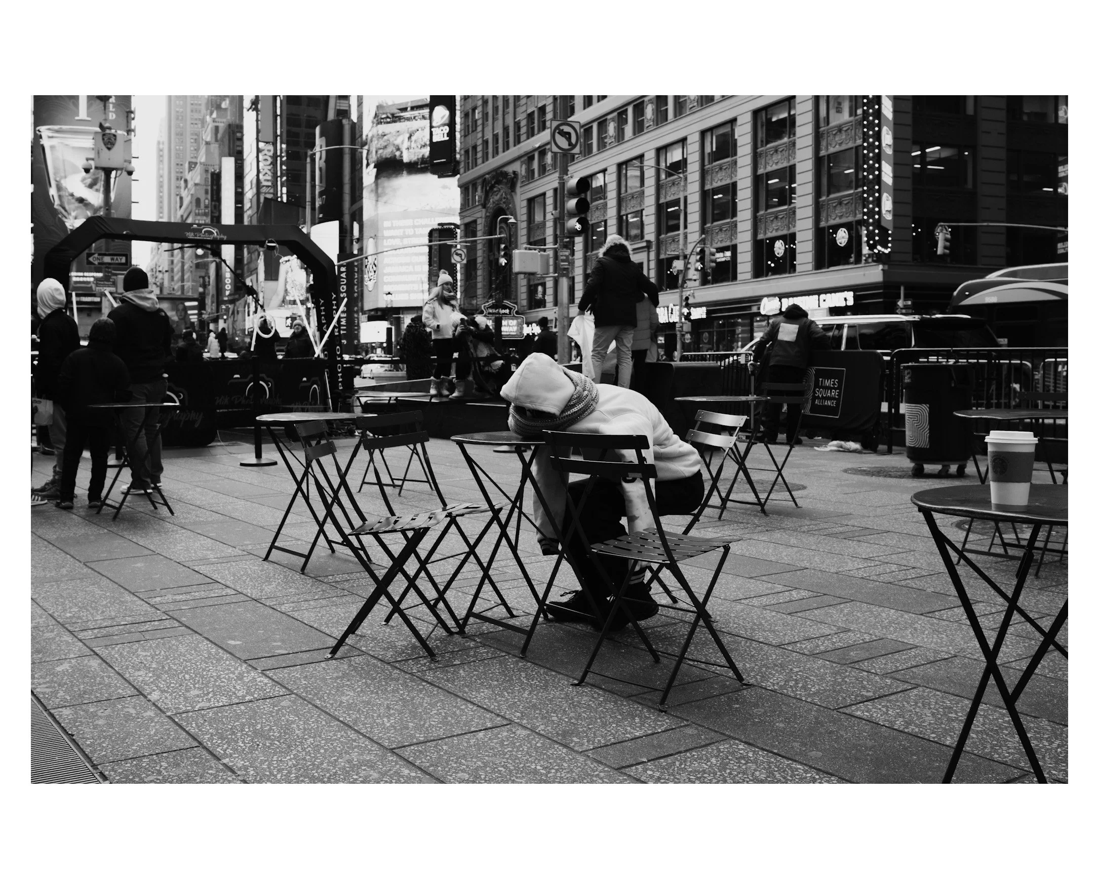 A black and white photo of Times Square showing outdoor seating with empty tables and chairs, a person sitting by themselves resting on a chair, and various people walking in the background amid tall buildings and bright digital billboards.