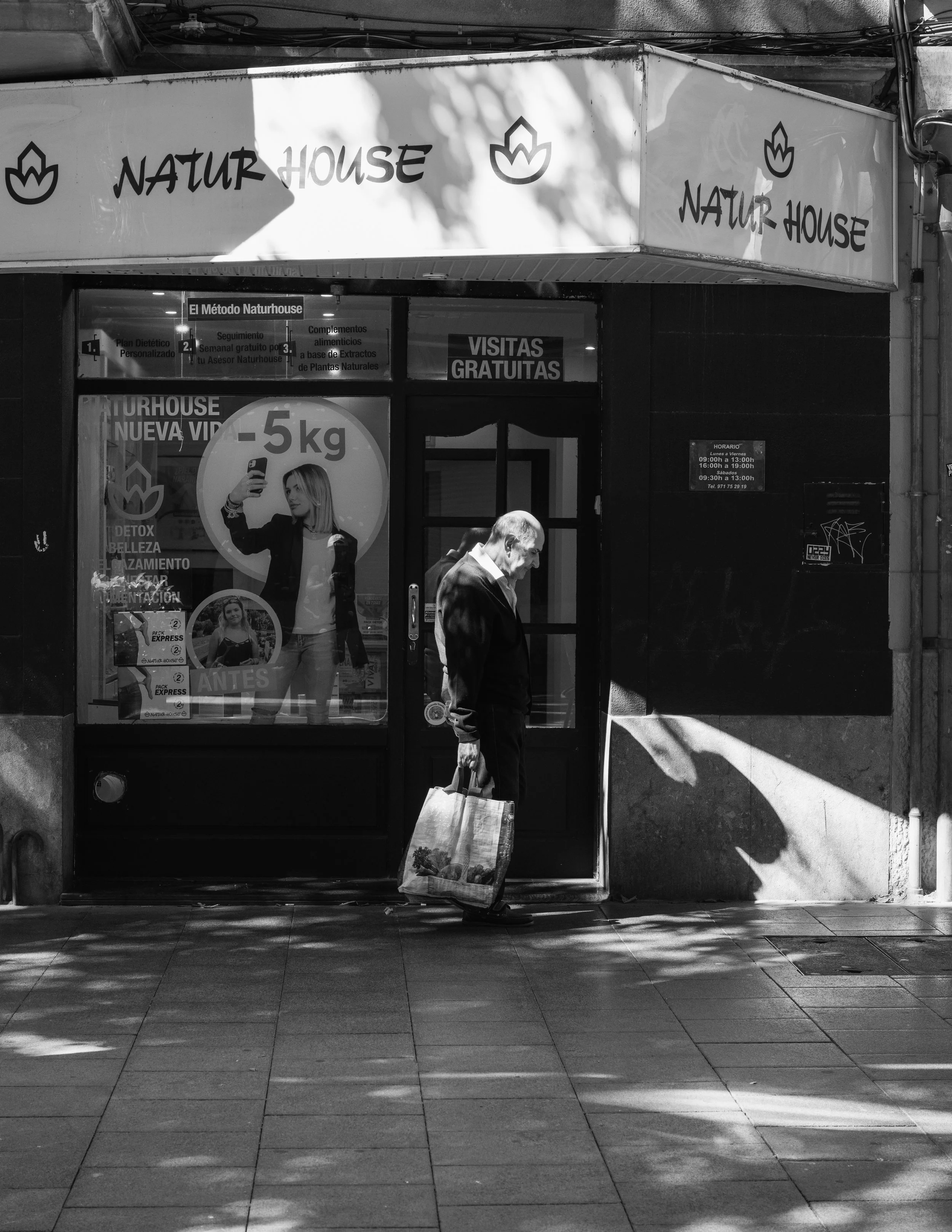 A black and white photo of a man with gray hair and a dark jacket walking past a store named Natur House. The man is carrying a tote bag with images on it. The store has a window with advertisements in Spanish, including a sign for free visits, and t