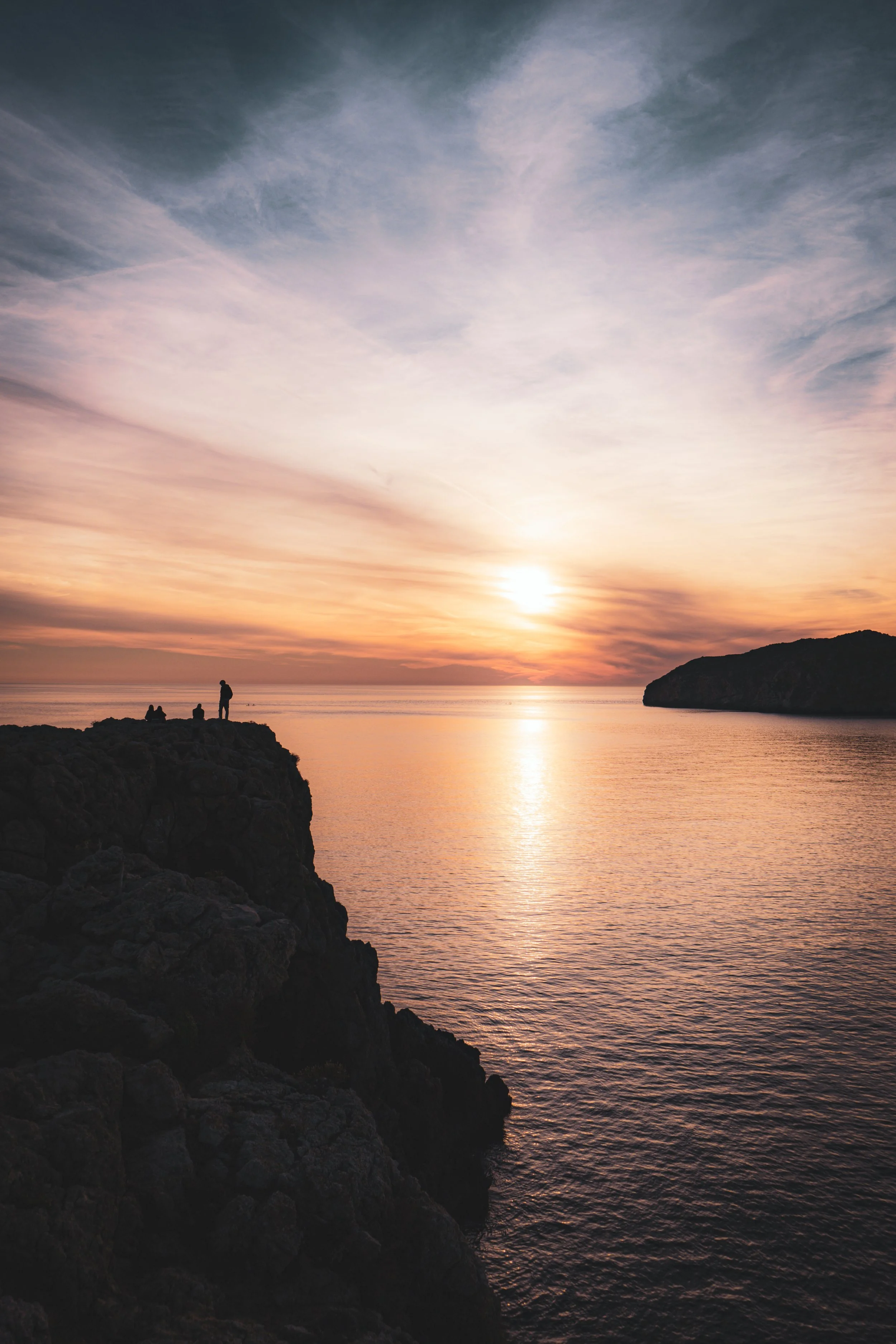 Silhouetted people on a rocky cliff watching a sunset over the ocean with a distant island and dramatic sky.