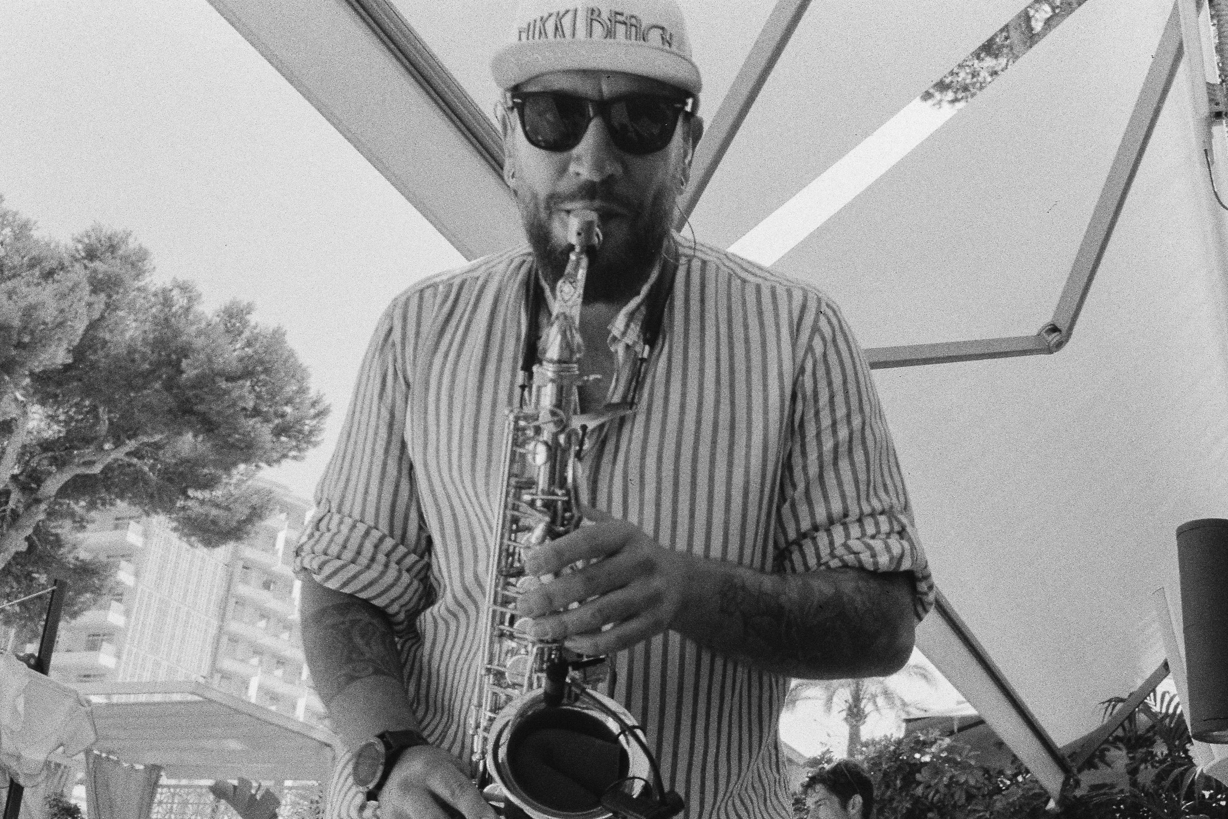 A man wearing sunglasses, a cap, and a striped shirt playing a saxophone outdoors, with trees and buildings in the background.