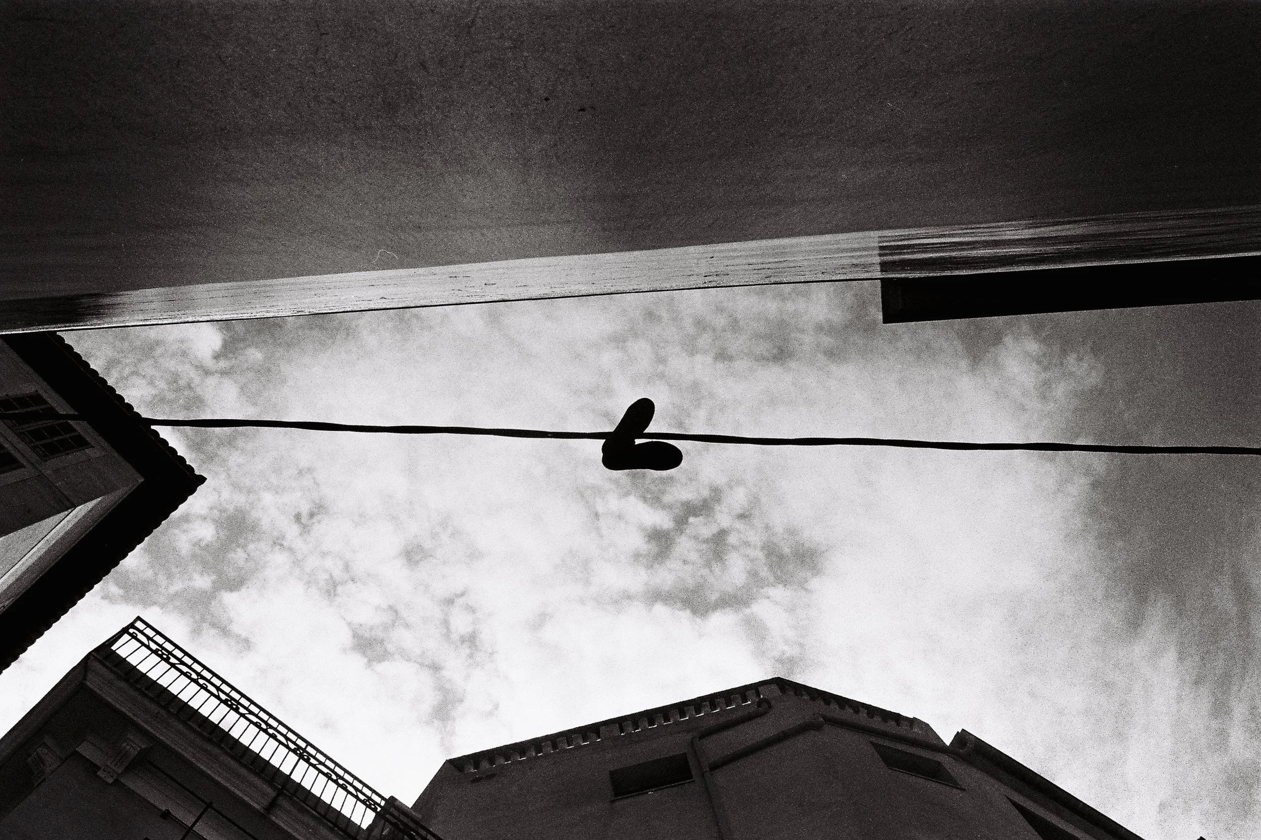 Black and white photograph of a sky viewed from below, with part of a building, a balcony, and a wire with a shoe hanging from it.