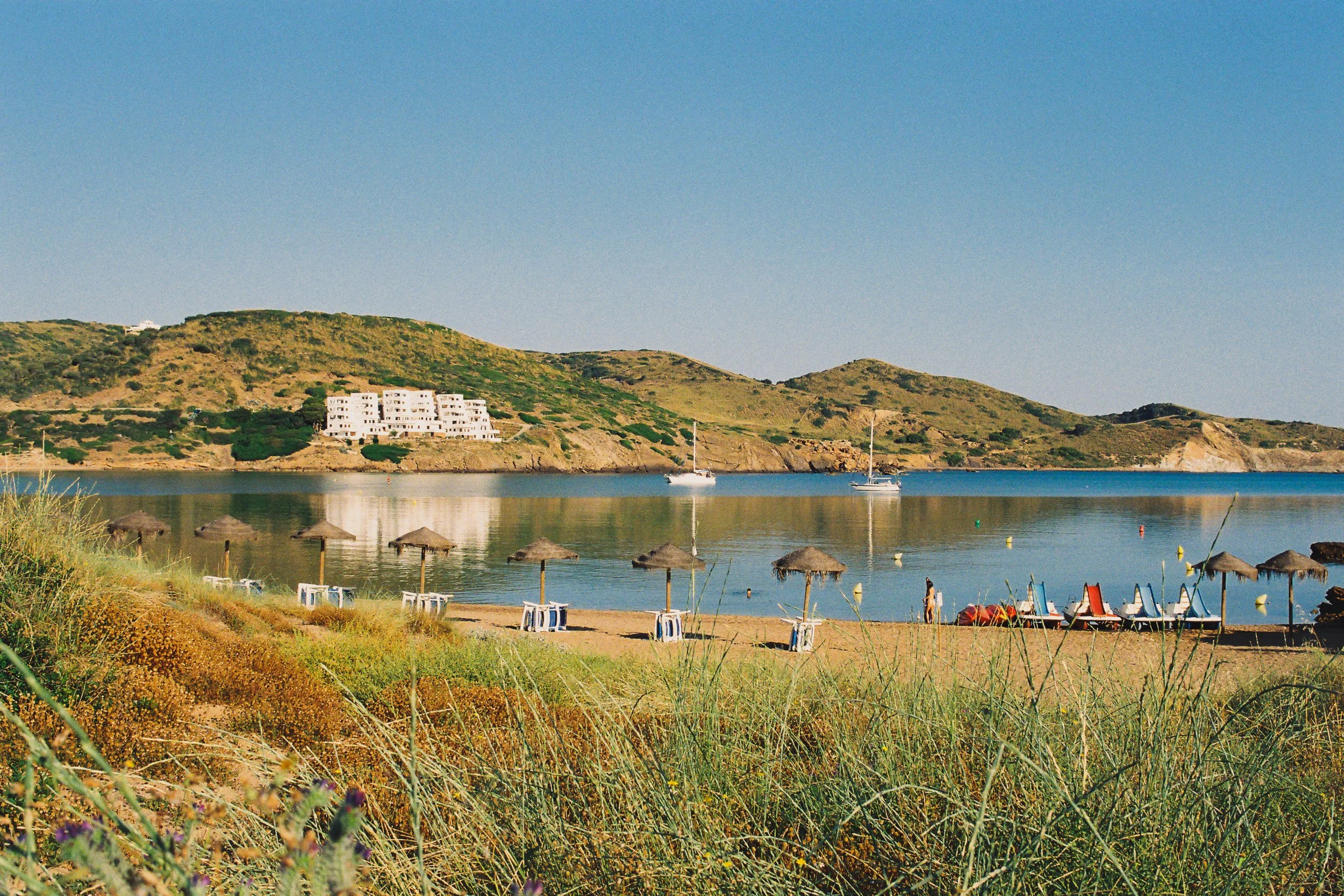 Beach scene with straw umbrellas, lounge chairs, and sailboats on calm water, with green hills and a white residential building in the background, under a clear blue sky.