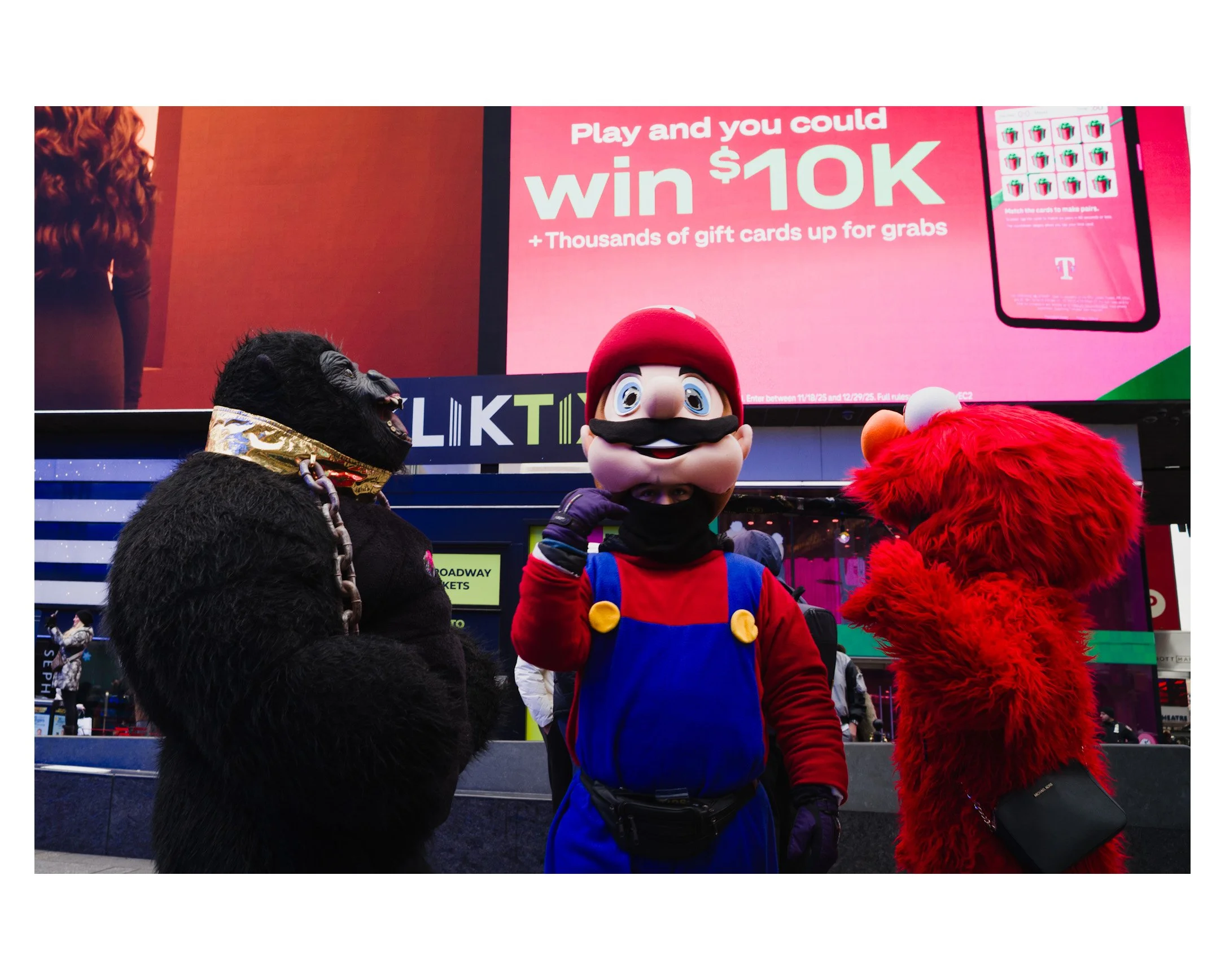 People dressed in costumes of a gorilla, Mario, and Elmo standing in Times Square, New York City, with a large pink billboard advertising a chance to win $10,000 and gift cards.